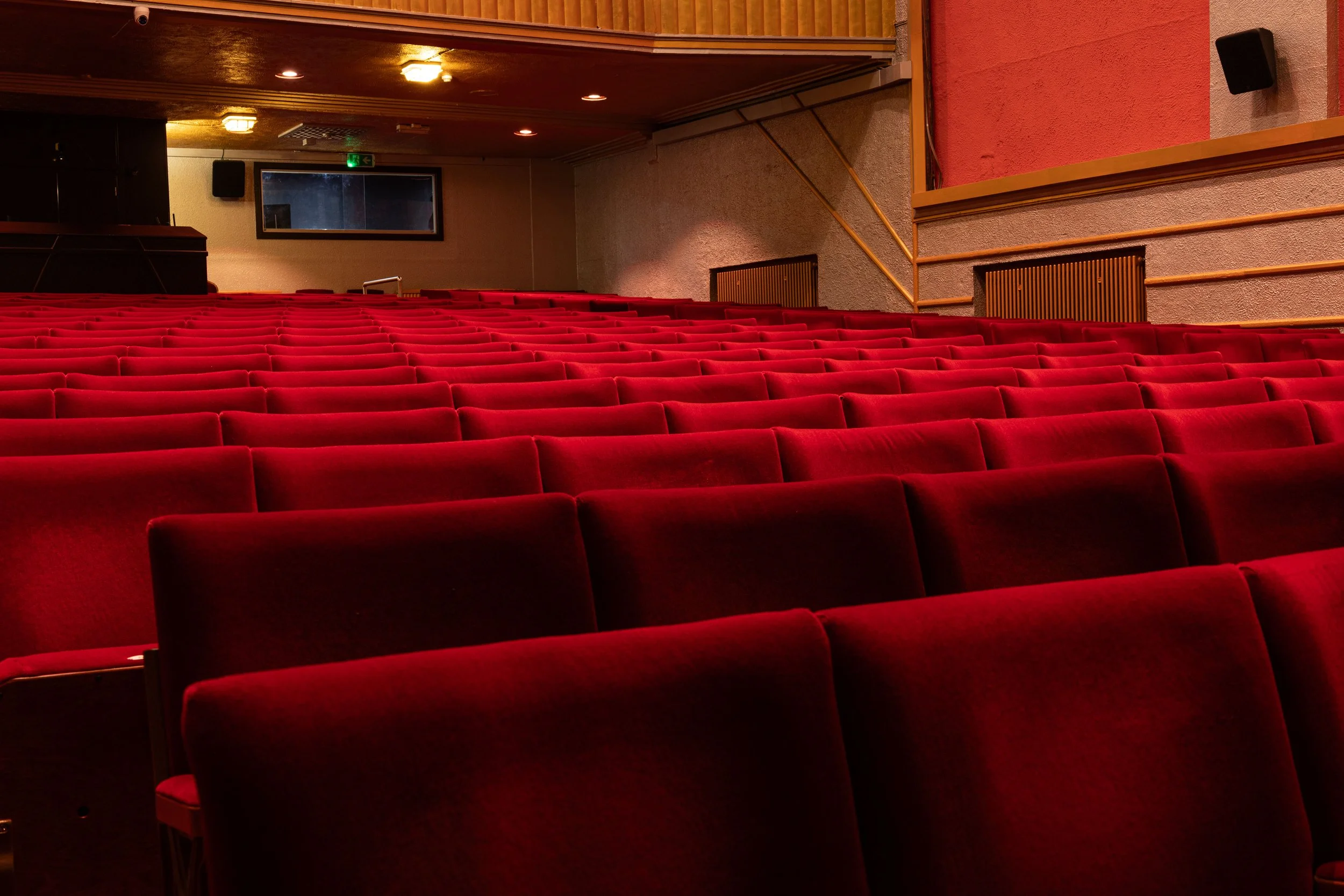 An empty theater auditorium with red velvet seats facing the stage. Walls are textured with beige and reddish paint, and there is a black speaker on the wall. A window and lighting fixtures are visible on the wall.