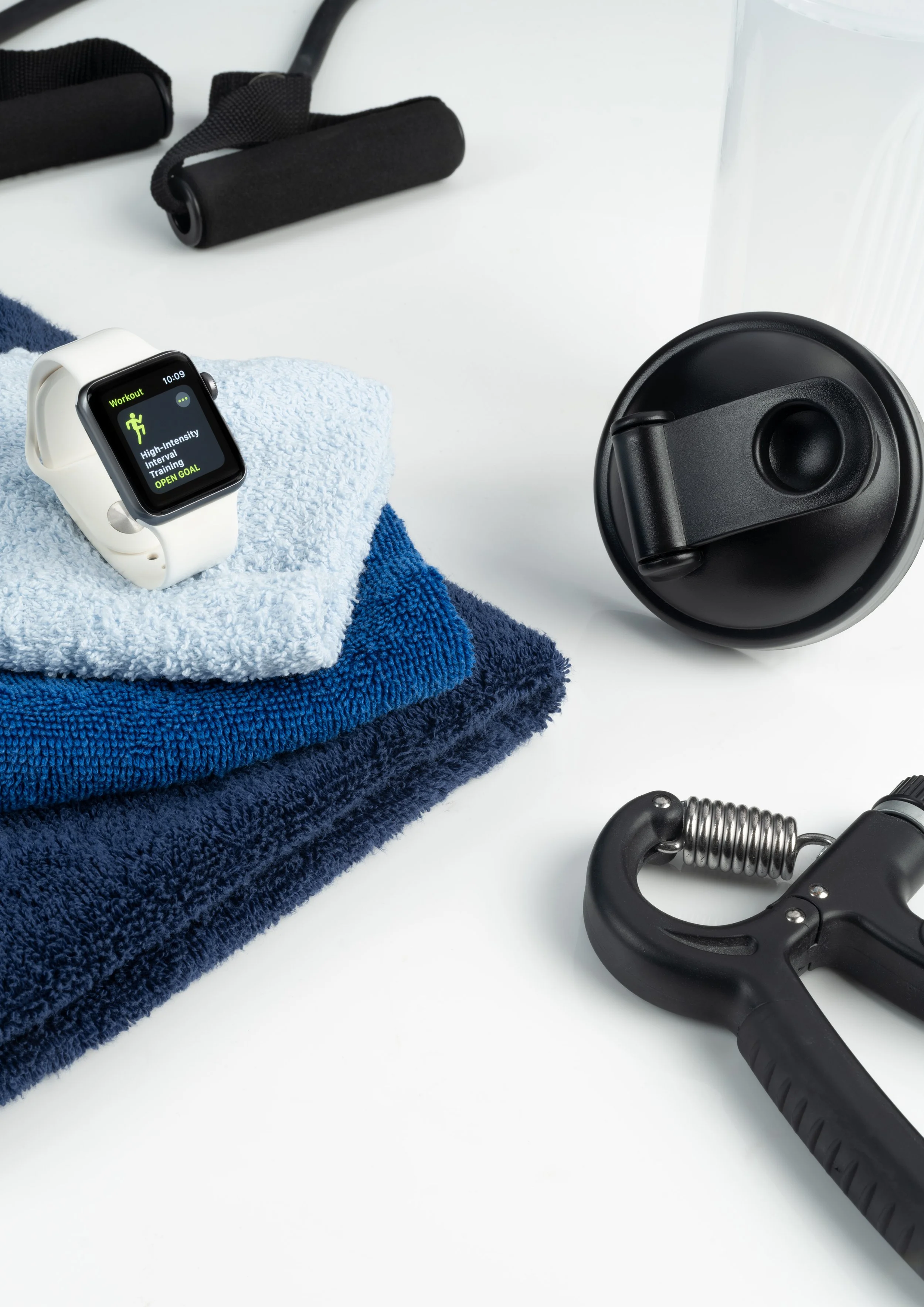 Fitness essentials including a smartwatch displaying a workout app, rolled blue towels, a watch, and a hand gripper on a white surface.