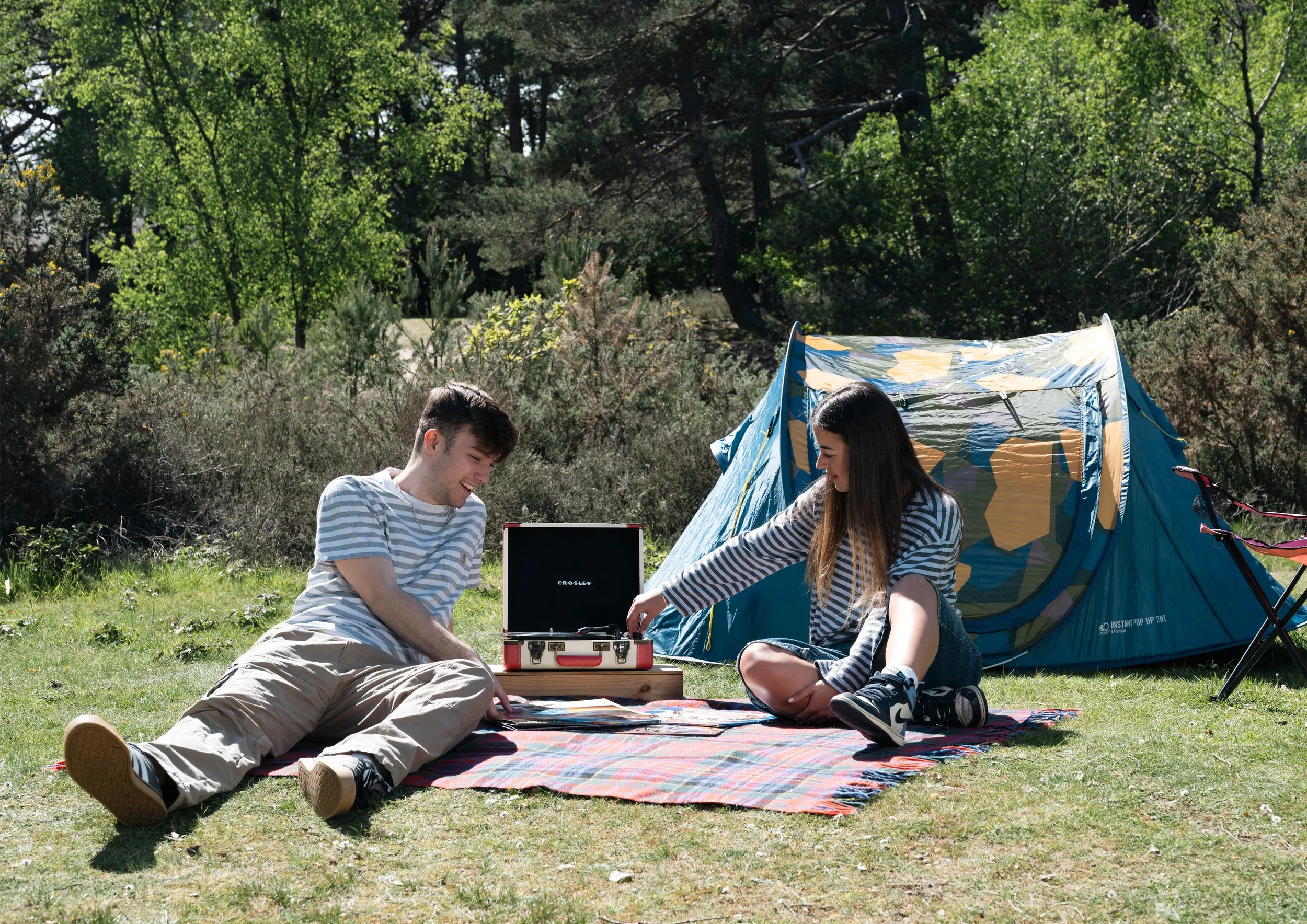 Two young people sitting on a colorful blanket at a campsite in the woods, with a blue and yellow tent nearby. They are smiling and looking at a laptop in a sunny outdoor setting with green trees in the background.