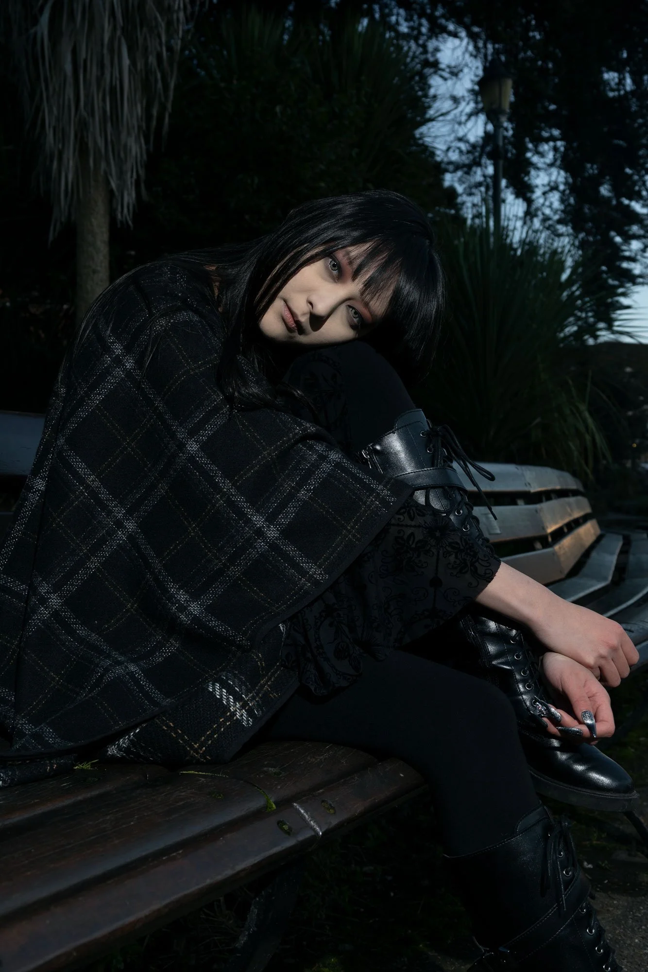 Young woman with black hair and pale skin sitting on a wooden park bench in a dark outdoor setting at dusk, wearing black boots and a black patterned outfit, with a background of trees and a streetlamp.