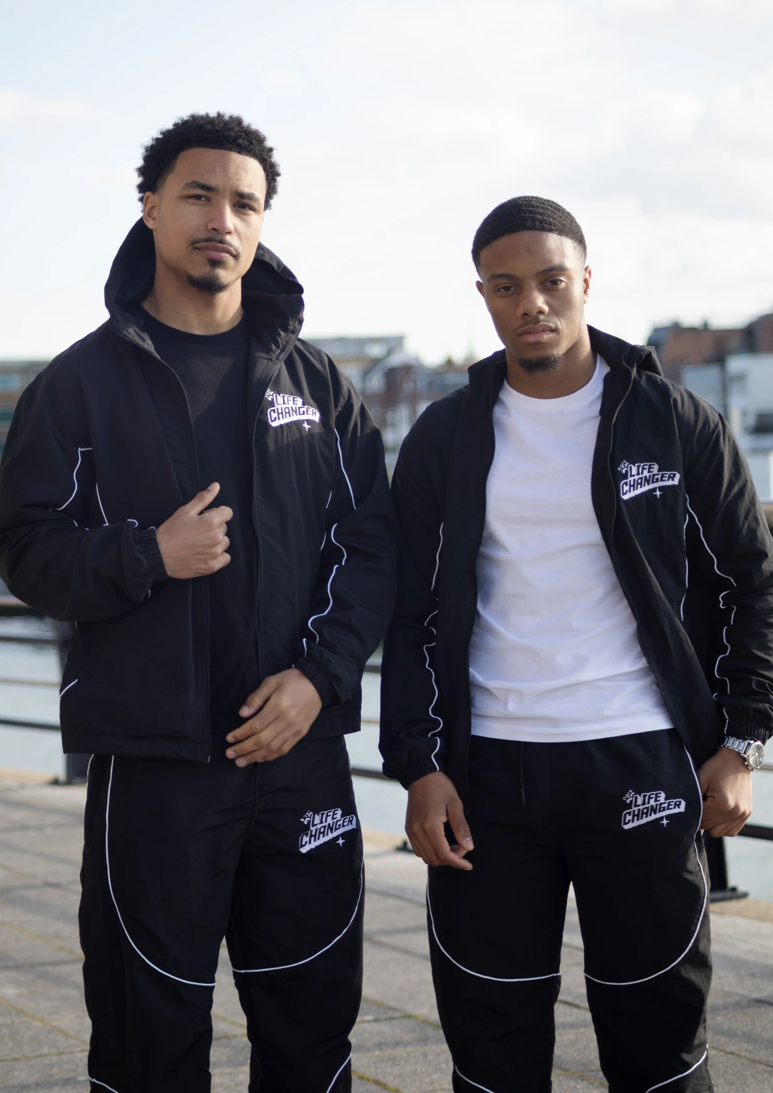 Two young men standing outdoors near a waterfront, wearing black jackets and pants with white accents and 'LIFE CHANGER' logos. They appear serious and confident.