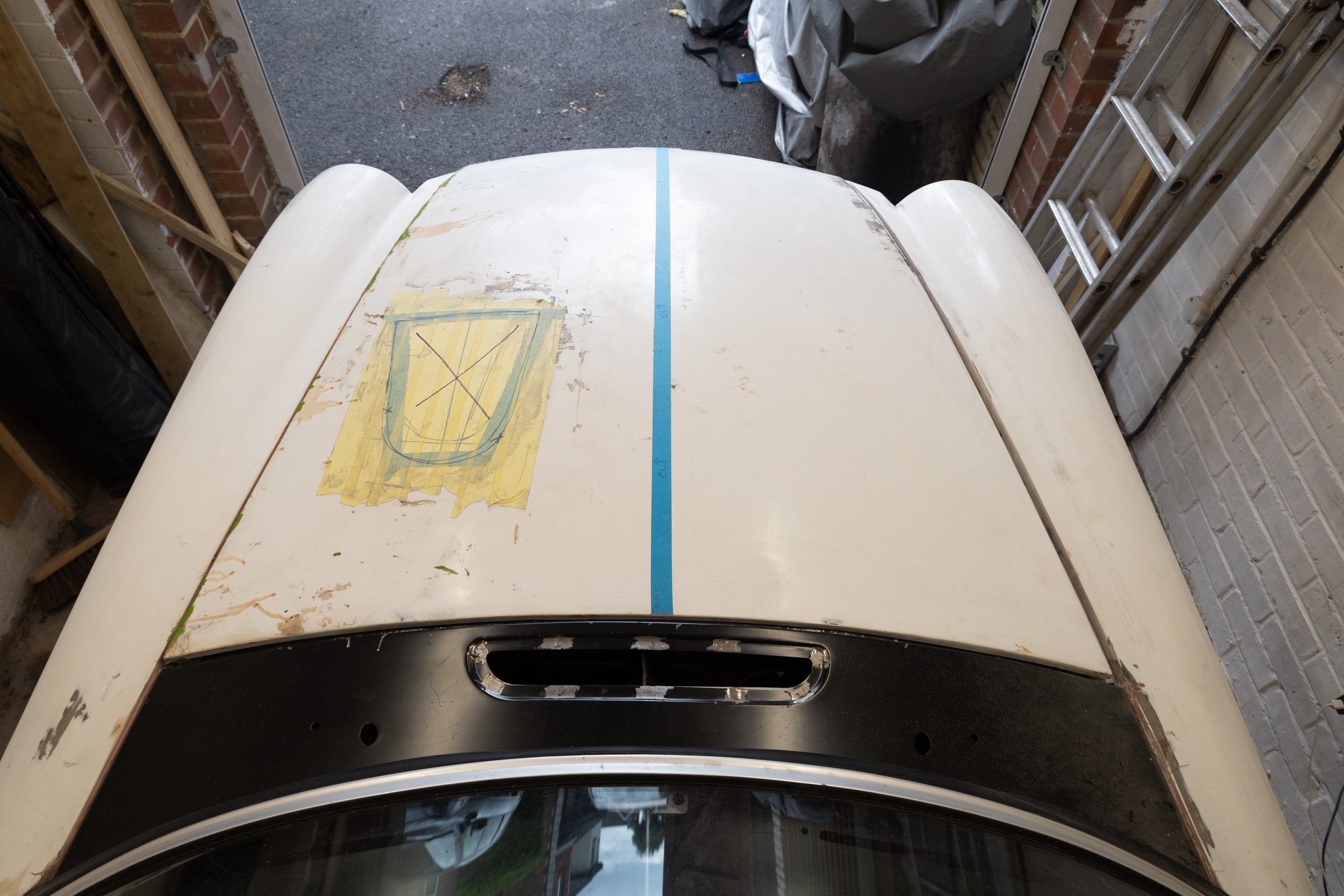 The image shows the top view of a white car with a partially painted surface, situated in a garage. There is a strip of blue painter's tape running down the center of the rear roof of the car, with some yellow painter's tape on the left side. The gar
