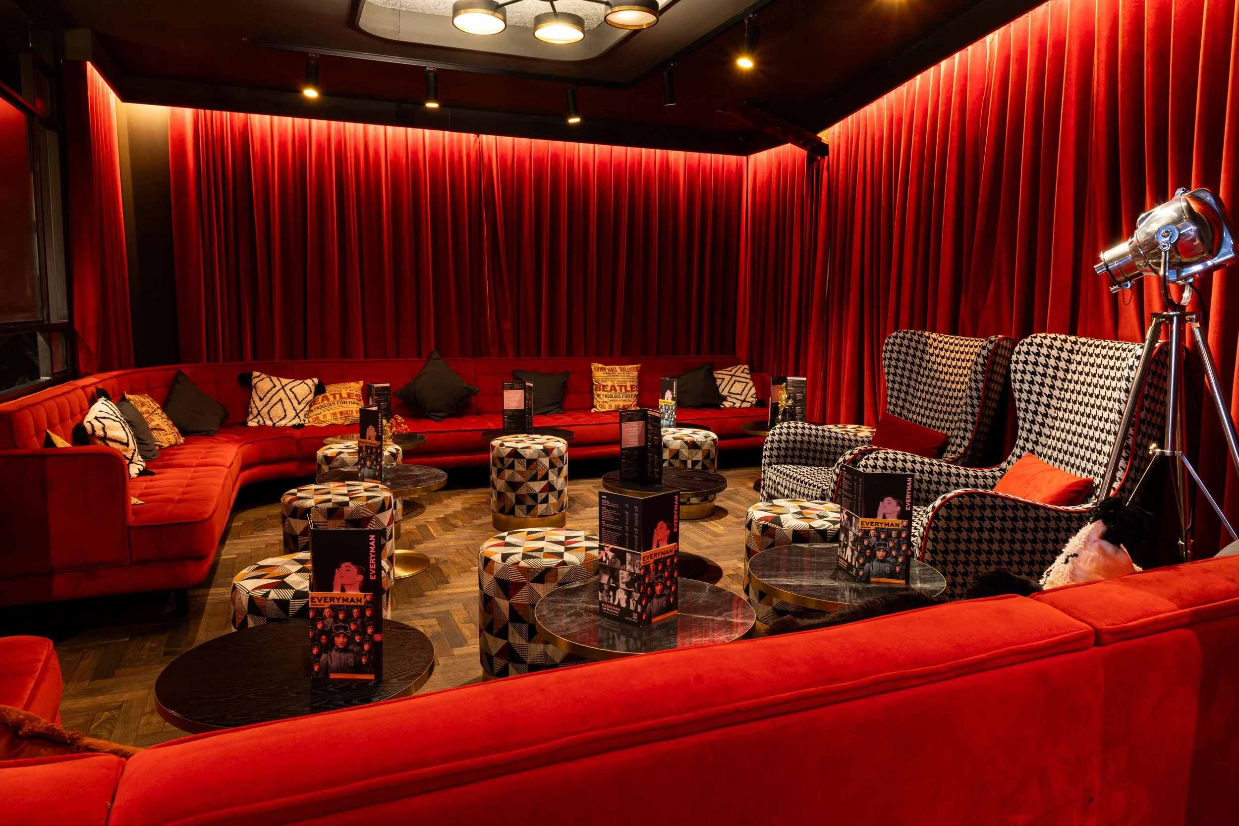 A cozy, stylish lounge area with red velvet curtains, a large red curved sofa, patterned armchairs, small round tables with magazines, and a vintage telescope on a tripod. Warm lighting enhances the inviting atmosphere.