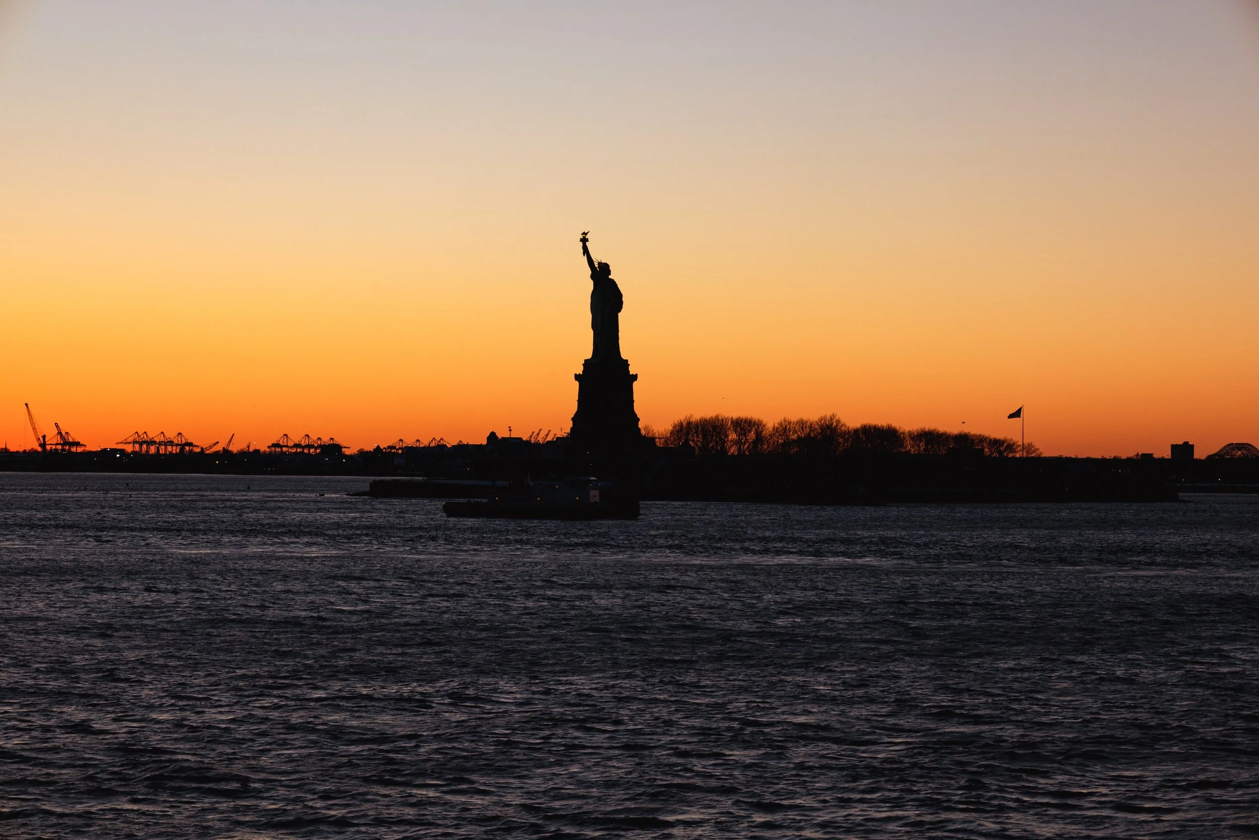 Silhouette of the Statue of Liberty against a colorful sunset sky with water in the foreground.