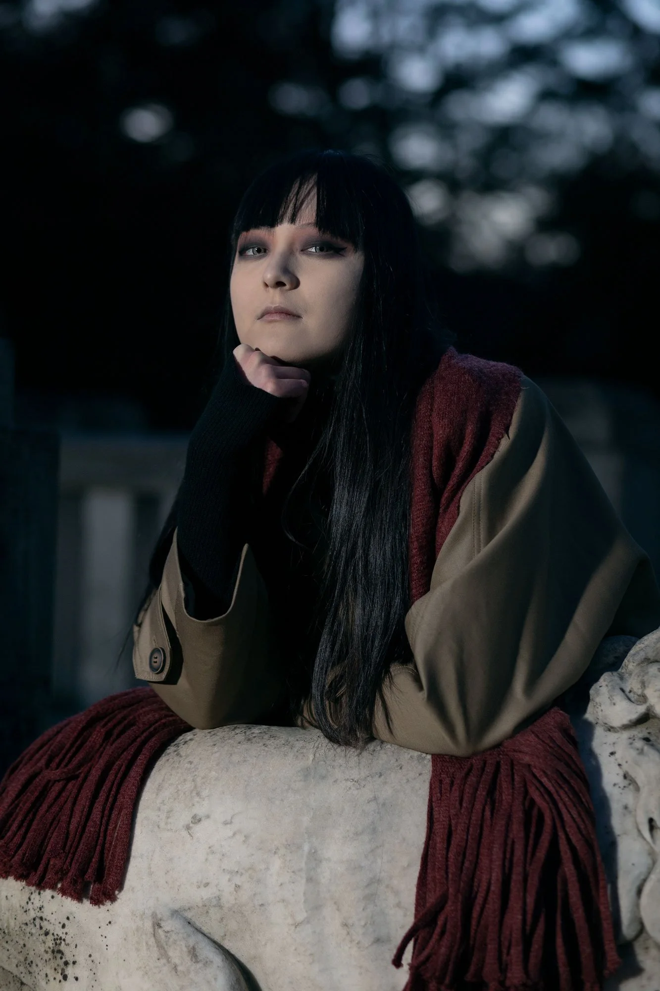 A woman with long dark hair and pale skin wearing makeup, a beige and red jacket, and a black glove resting her chin on her hand, sitting outdoors on a large stone with snow and trees in the background during dusk.