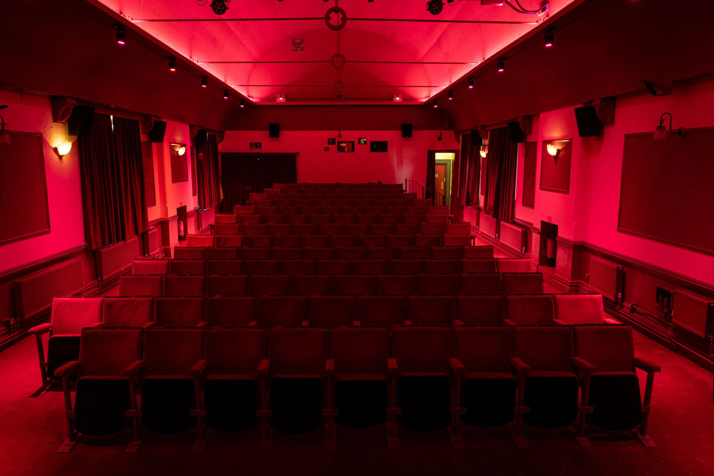 Empty theater with red lighting, rows of red seats, curtains, and wall-mounted lights.