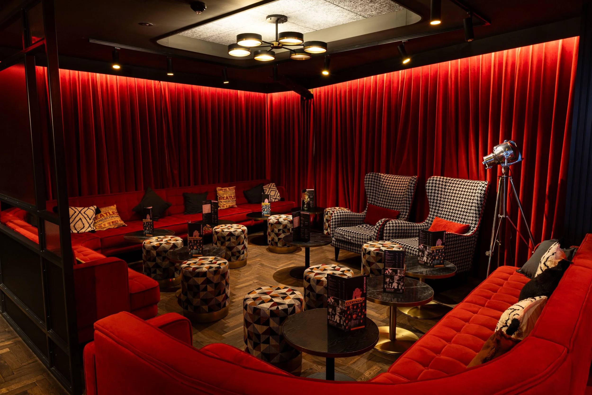 cozy lounge with red velvet sofa and houndstooth patterned armchairs, small round tables, black and white pillows, red curtains, and modern overhead lighting