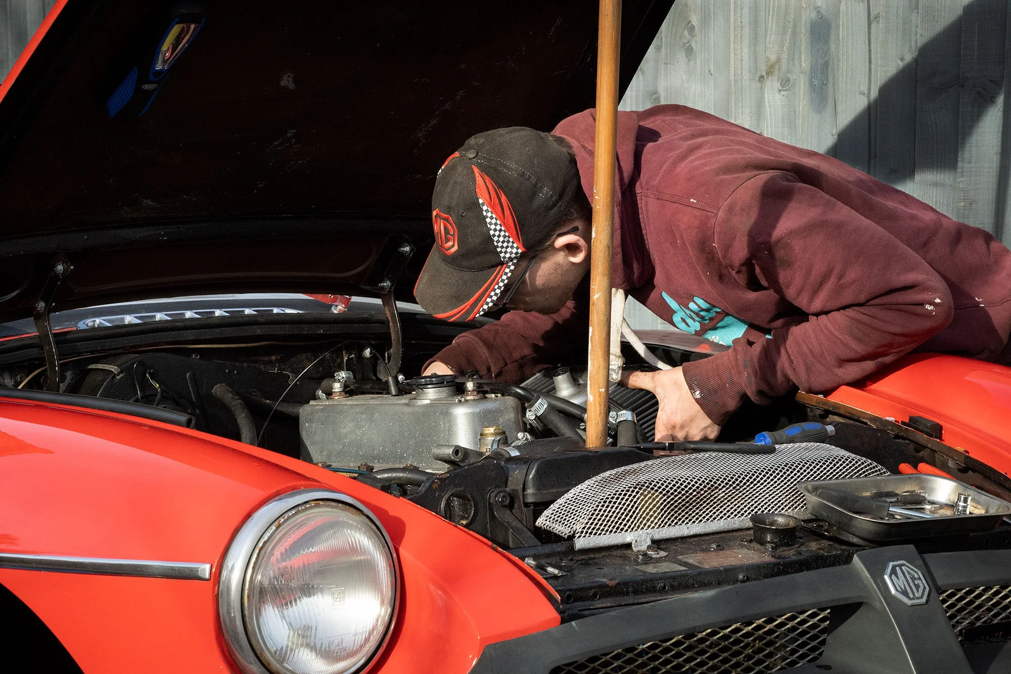 A person working under the hood of a red vintage MG car, wearing a black cap with orange and white details and a maroon hoodie, at an outdoor location.