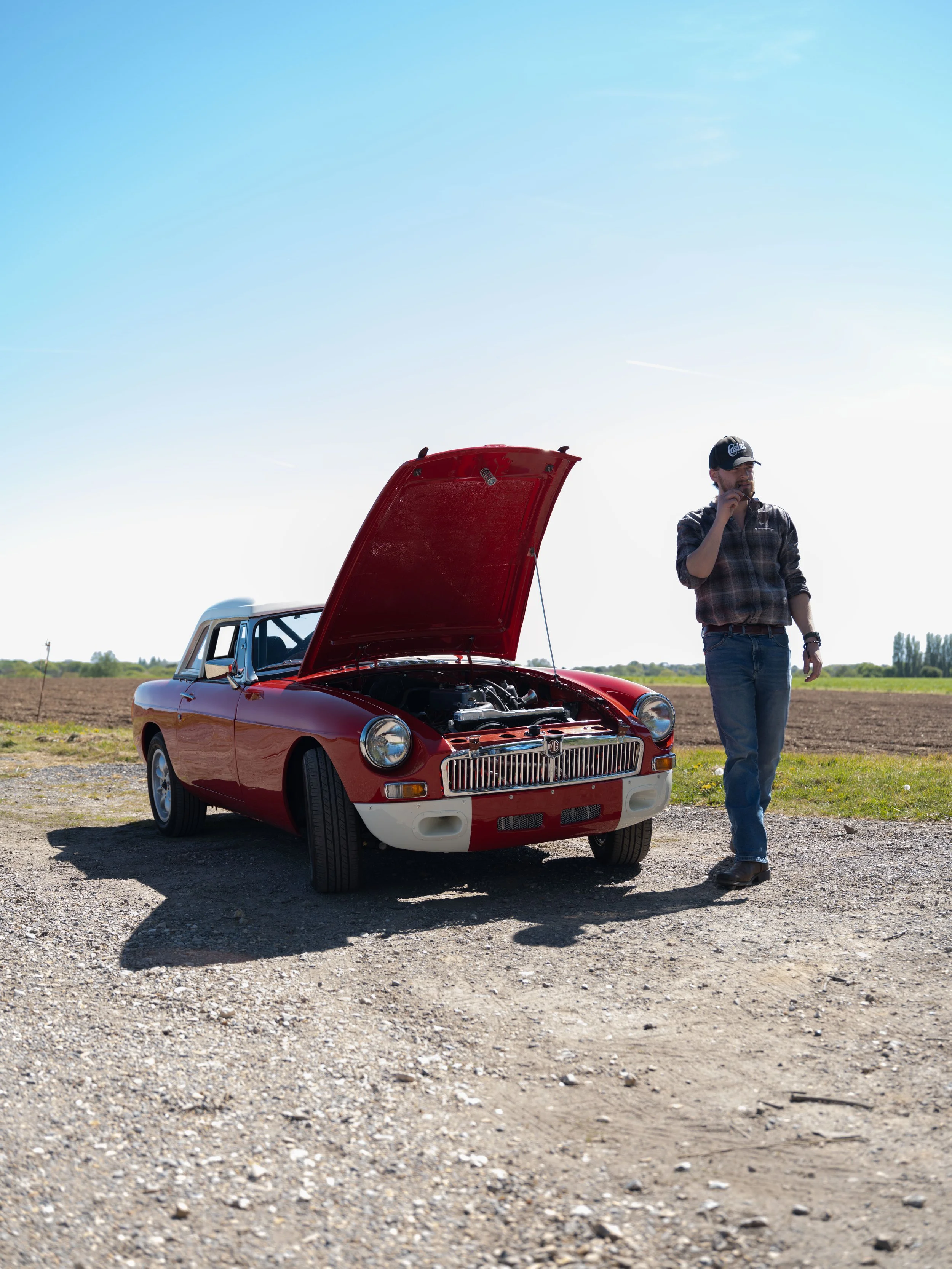A man standing next to a red vintage car with its hood open on a dirt patch in a rural area on a sunny day.