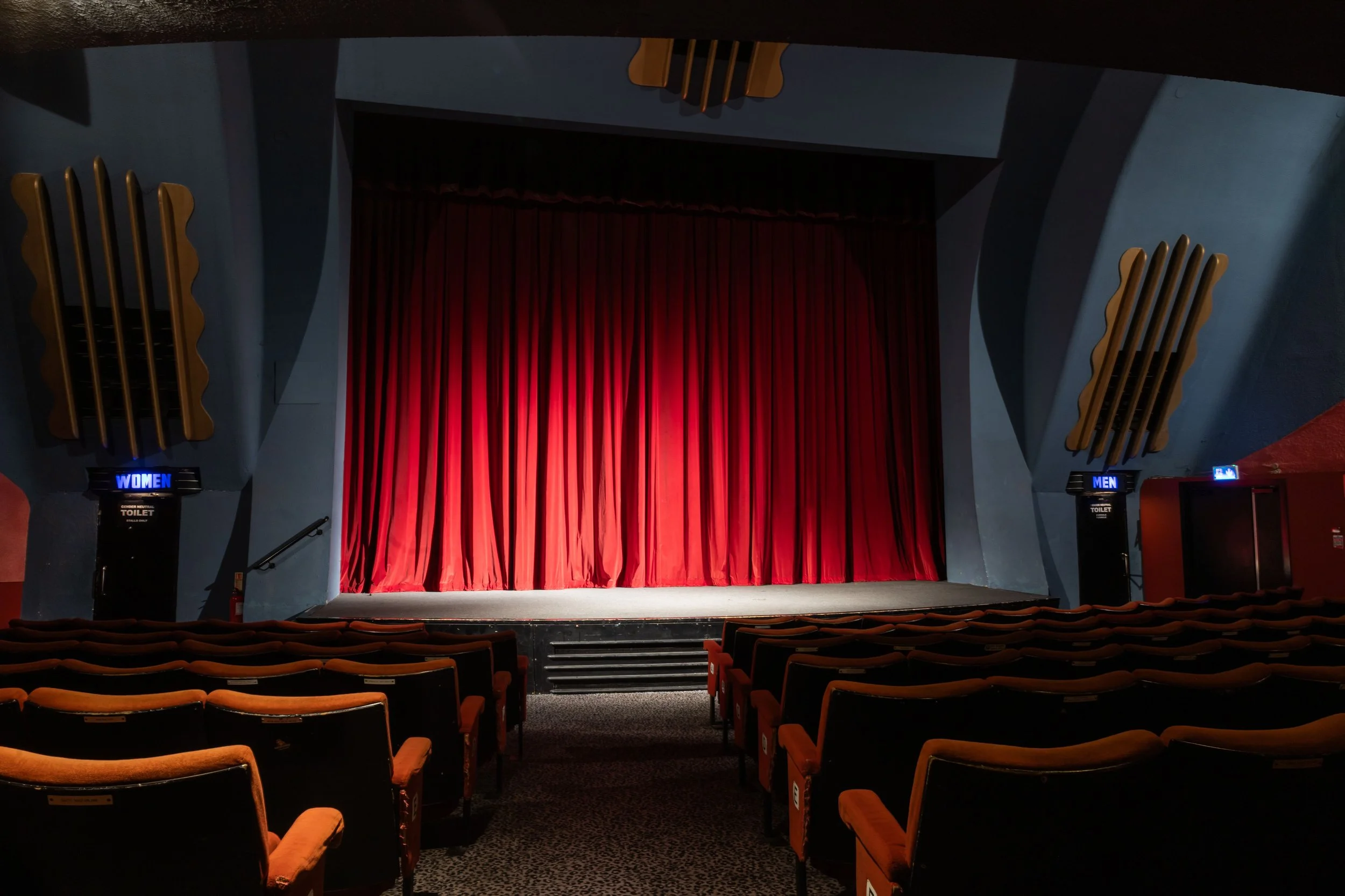 The interior of a theater with plush orange seats, a stage with a red curtain, and directional signs for women's and men's restrooms.