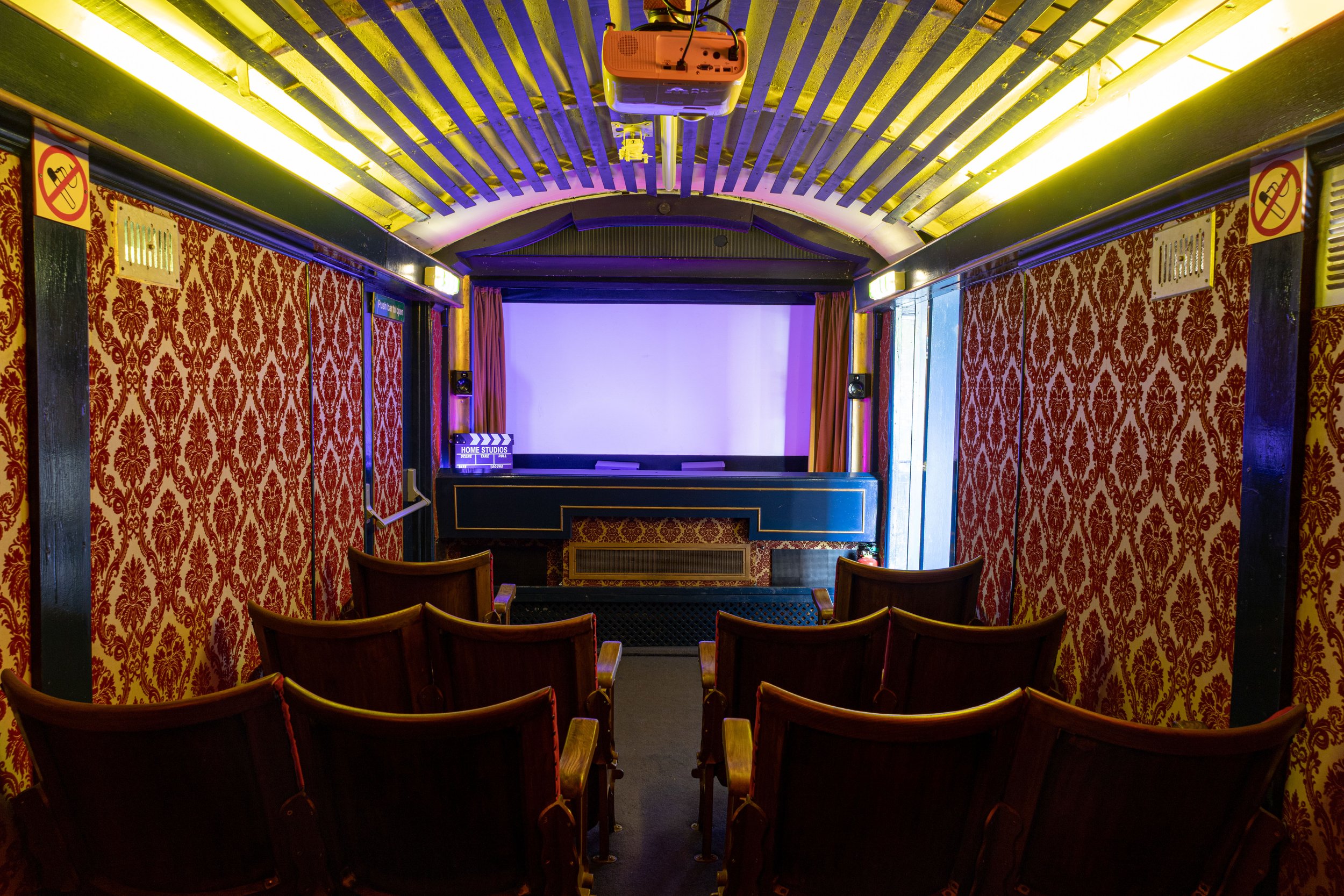 An empty small theater or screening room with red damask wallpaper, wooden chairs arranged in rows, a large white screen, and a projector mounted on the ceiling.