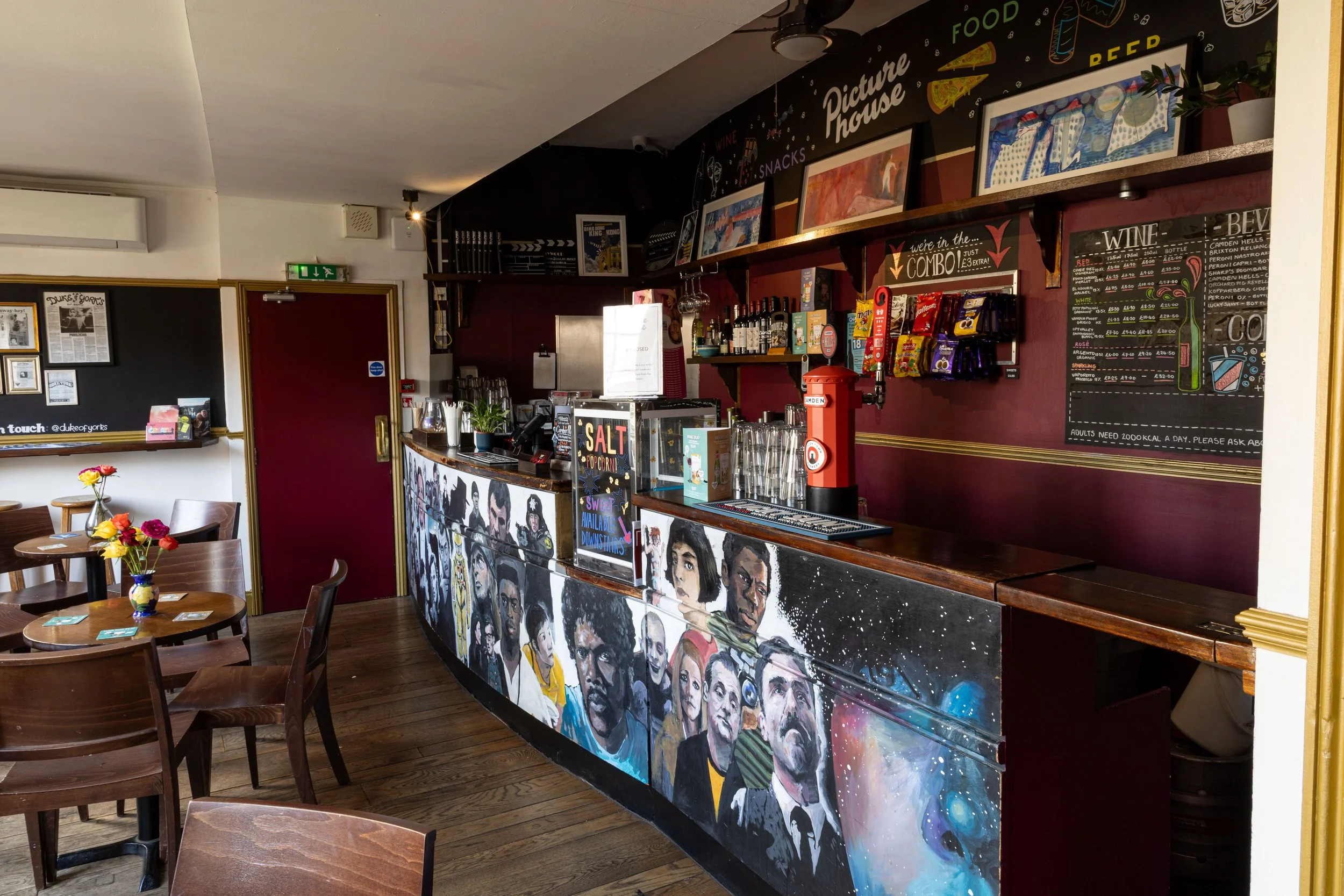 Interior of a cafe with a colorful painted front desk featuring portraits of diverse people. The cafe has wooden tables with small vases of flowers, and a menu board displaying wine, beer, and cocktails. A blackboard on the wall lists food, snacks, a