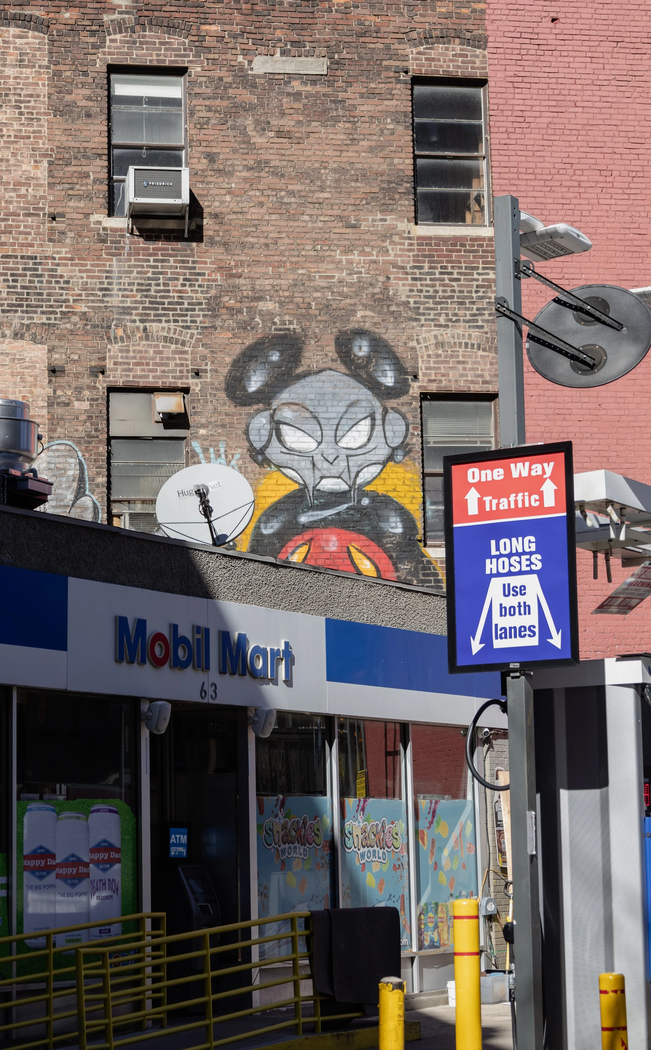 Graffiti of a stylized alien face with large eyes and antennae, painted on a brick wall above the entrance of a Mobil Mart store. The store has yellow metal barriers outside, a satellite dish, and a street sign indicating one-way traffic and lane usa