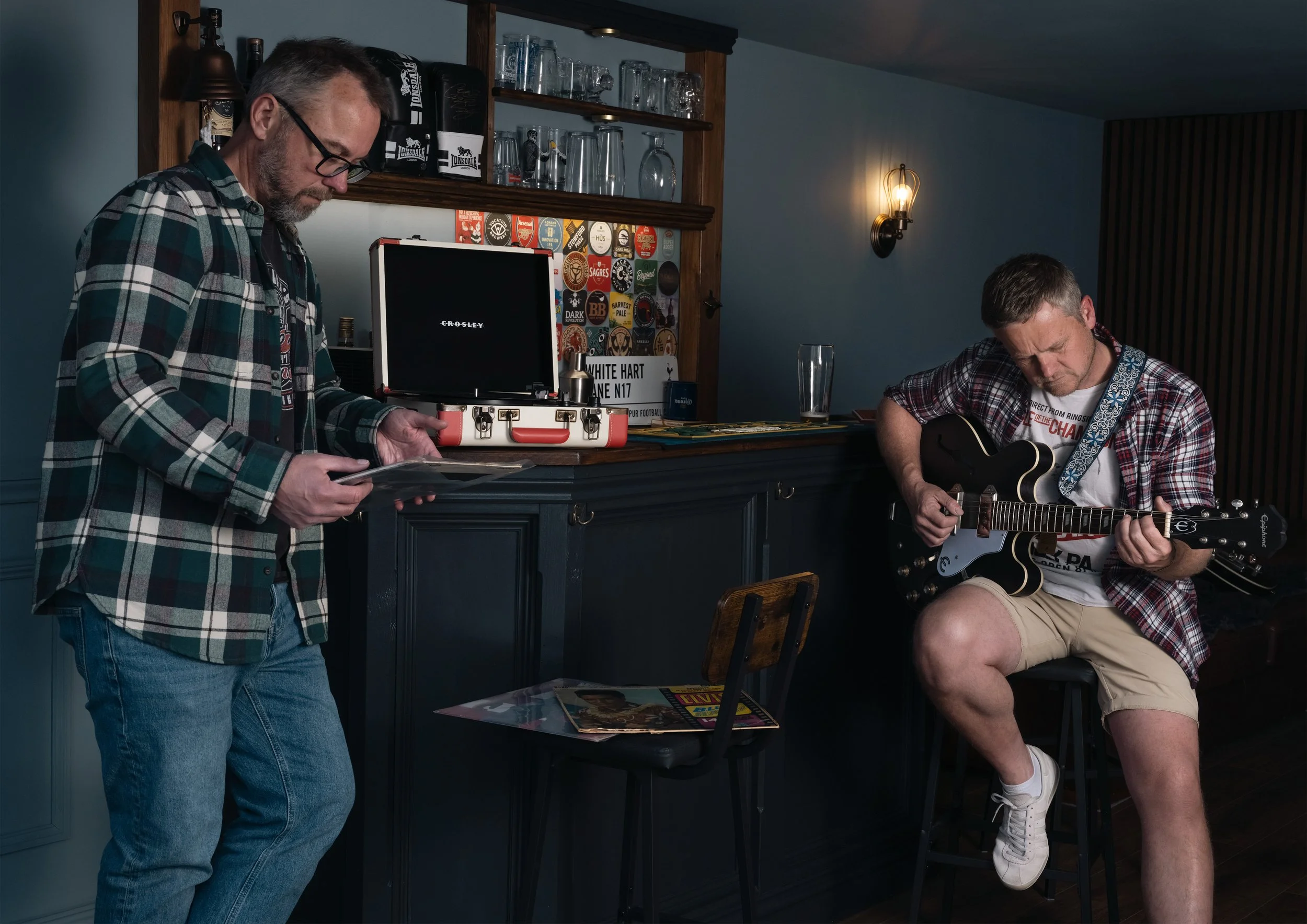 Two men in casual clothing, one standing and one sitting, playing musical instruments in a bar or pub setting. The man on the left holds a smartphone, and the man on the right is playing an electric guitar. There are glasses and beer coasters on a sh
