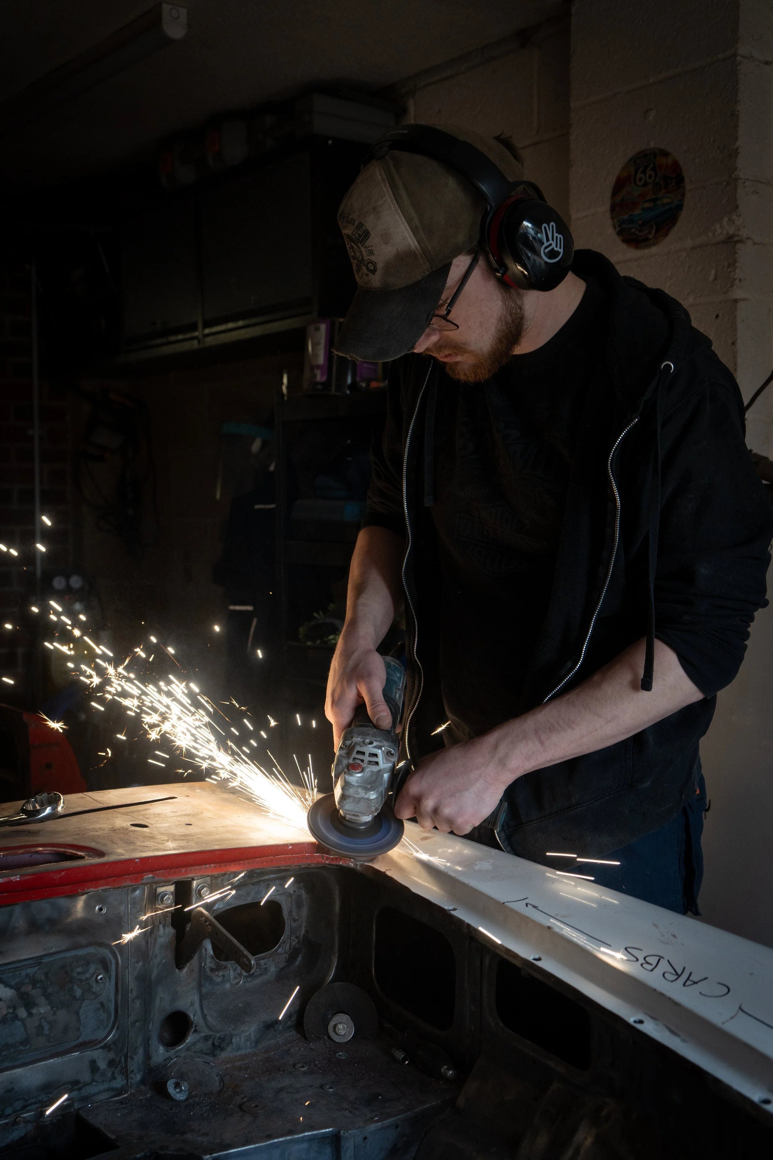 A man wearing a baseball cap, glasses, and noise-canceling headphones is using an angle grinder to cut or grind a piece of metal inside a car frame. Sparks are flying from the metal work, and the person is focused on the task. The setting appears to 