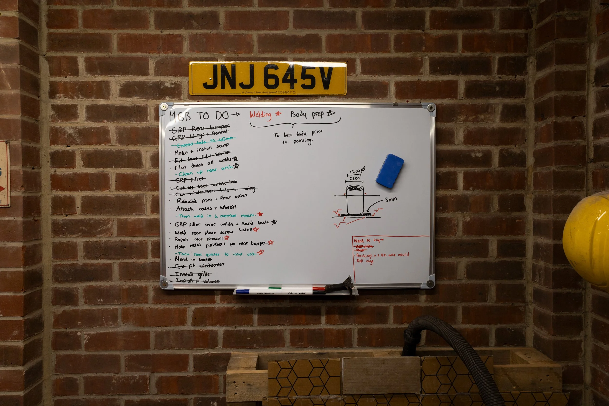 A brick wall with a yellow license plate reading 'JNJ 645V' mounted above a whiteboard. The whiteboard contains handwritten notes and diagrams related to a welding and bodywork project, with a blue eraser attached to its top edge. There is a yellow b
