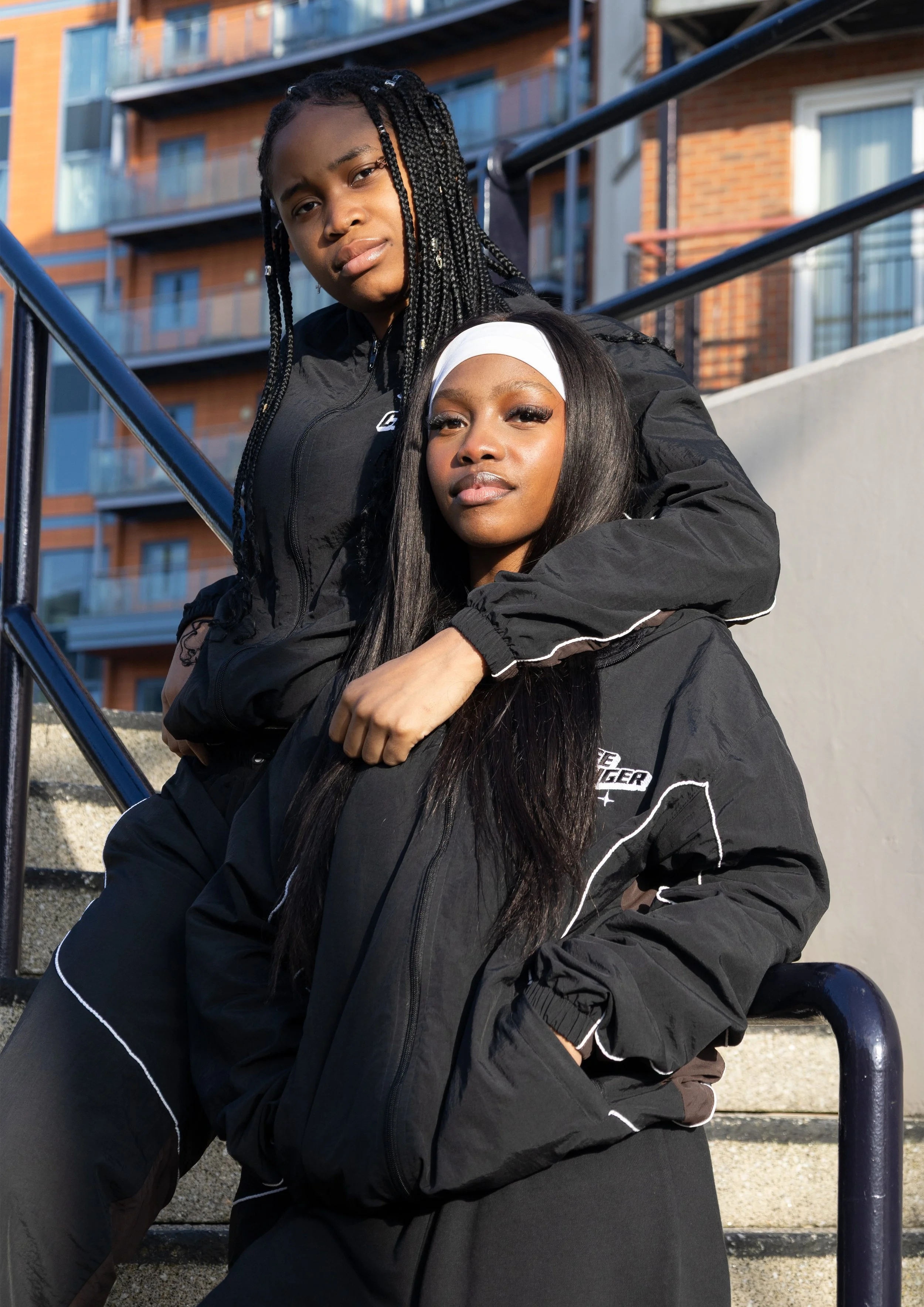 Two young women with black hair styled in braids and straight hair, wearing black jackets, sitting on outdoor stairs with a modern apartment building in background.