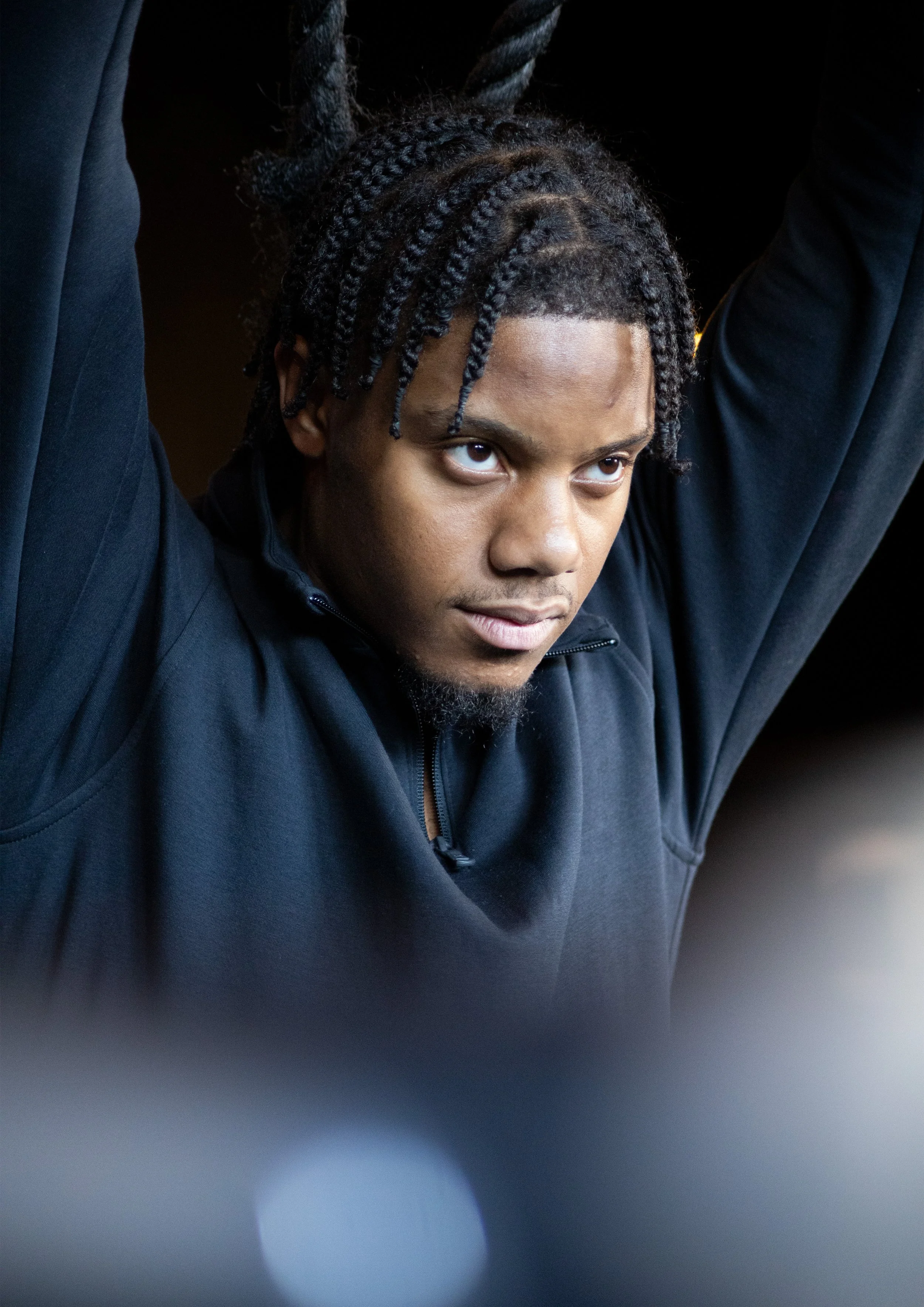 A person with braided hair tied into two high puffs, wearing a black zip-up jacket, looking downward with arms raised, in a dark setting.