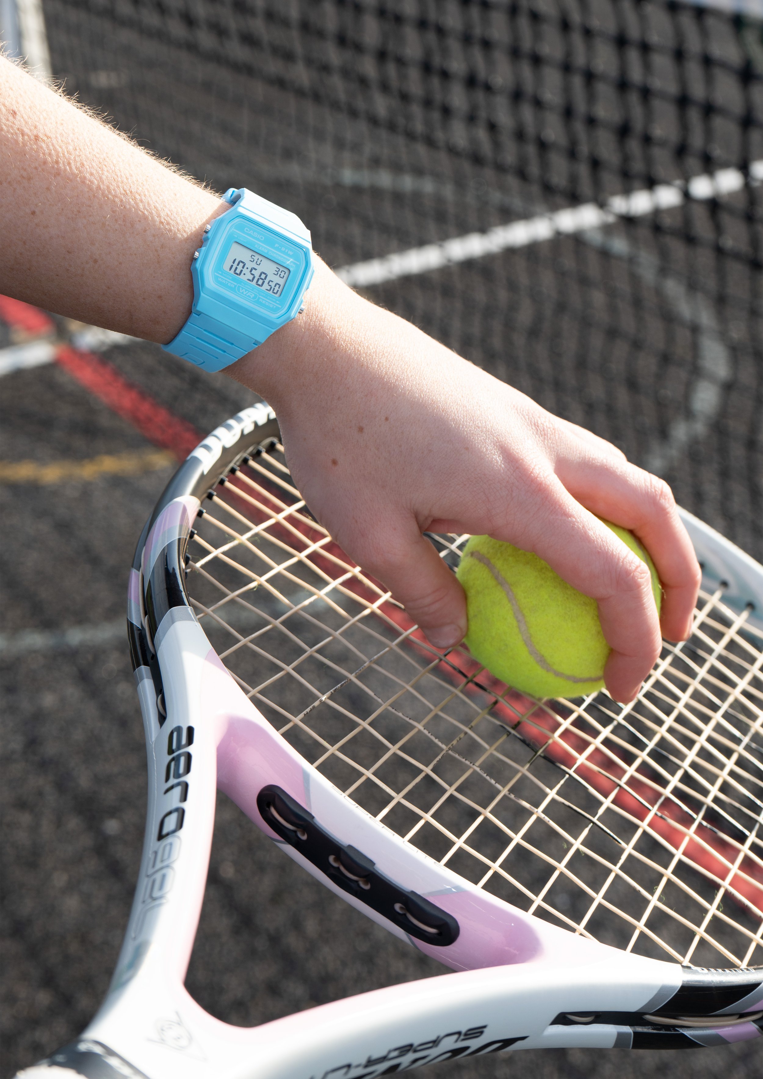 A person with a blue digital watch on their wrist, holding a yellow tennis ball, with a tennis racket on a tennis court.