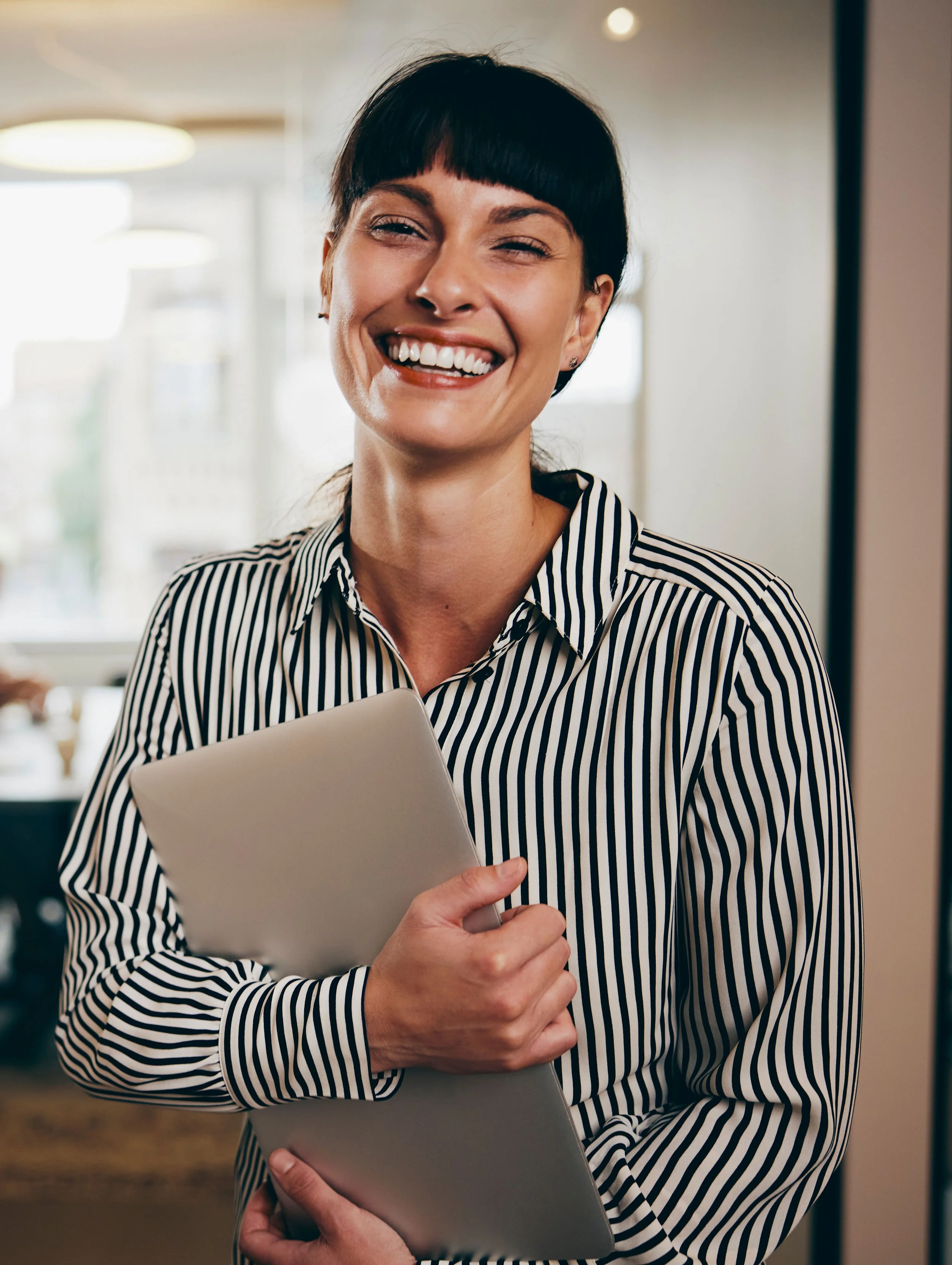 A woman with short dark hair, wearing a black and white striped blouse, holding a silver laptop, smiling showing her teeth, in an indoor setting with blurred background lighting.