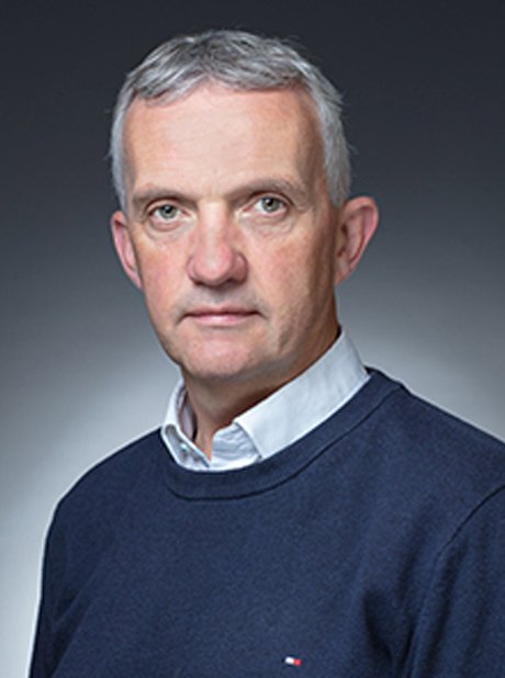 Portrait of a middle-aged man with short gray hair, wearing a white collared shirt and a dark blue sweater, against a gray background.