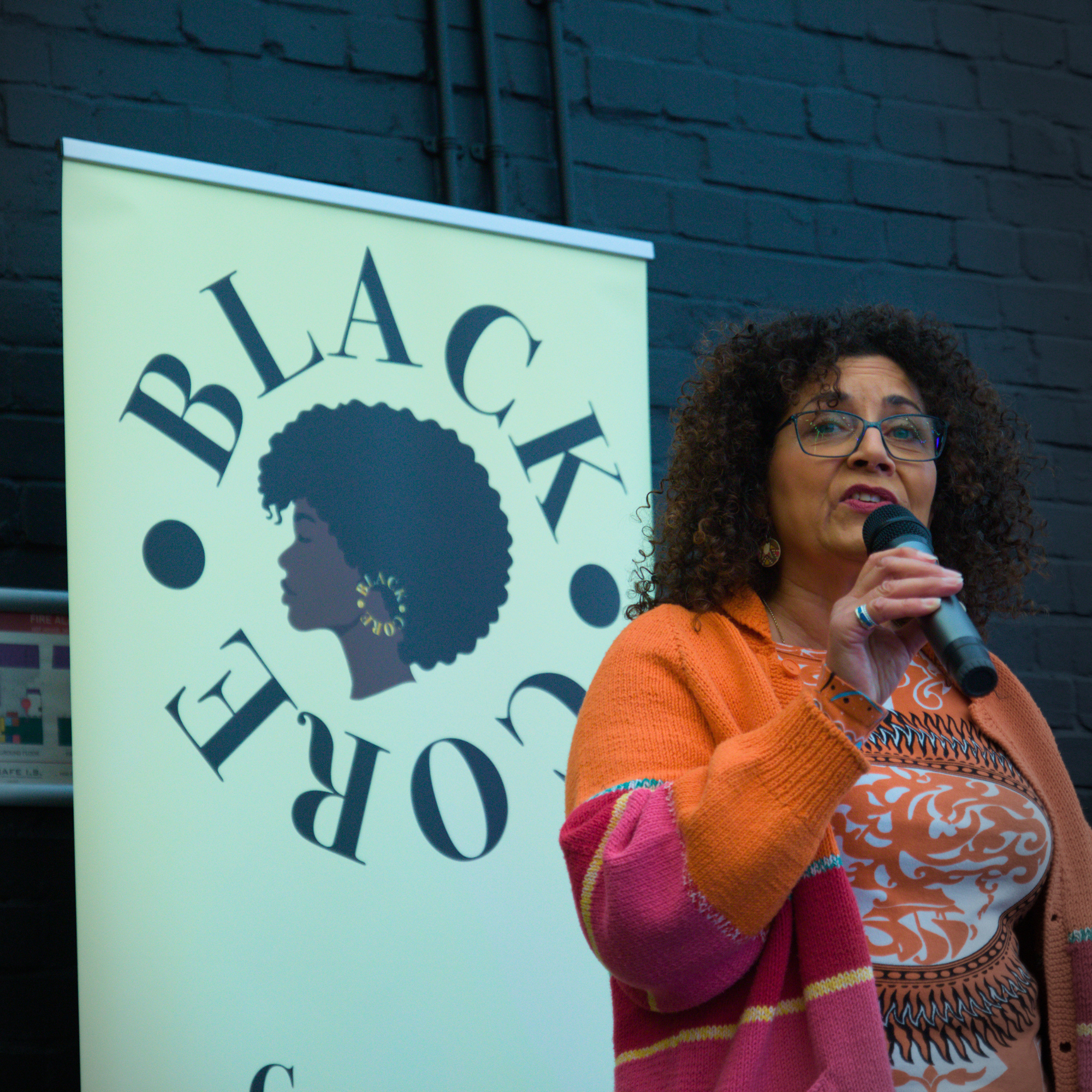 A woman with curly hair and glasses speaking into a microphone at an indoor event, standing next to a yellow poster with black text and an illustration of a woman's silhouette with natural hair, against a dark brick wall.