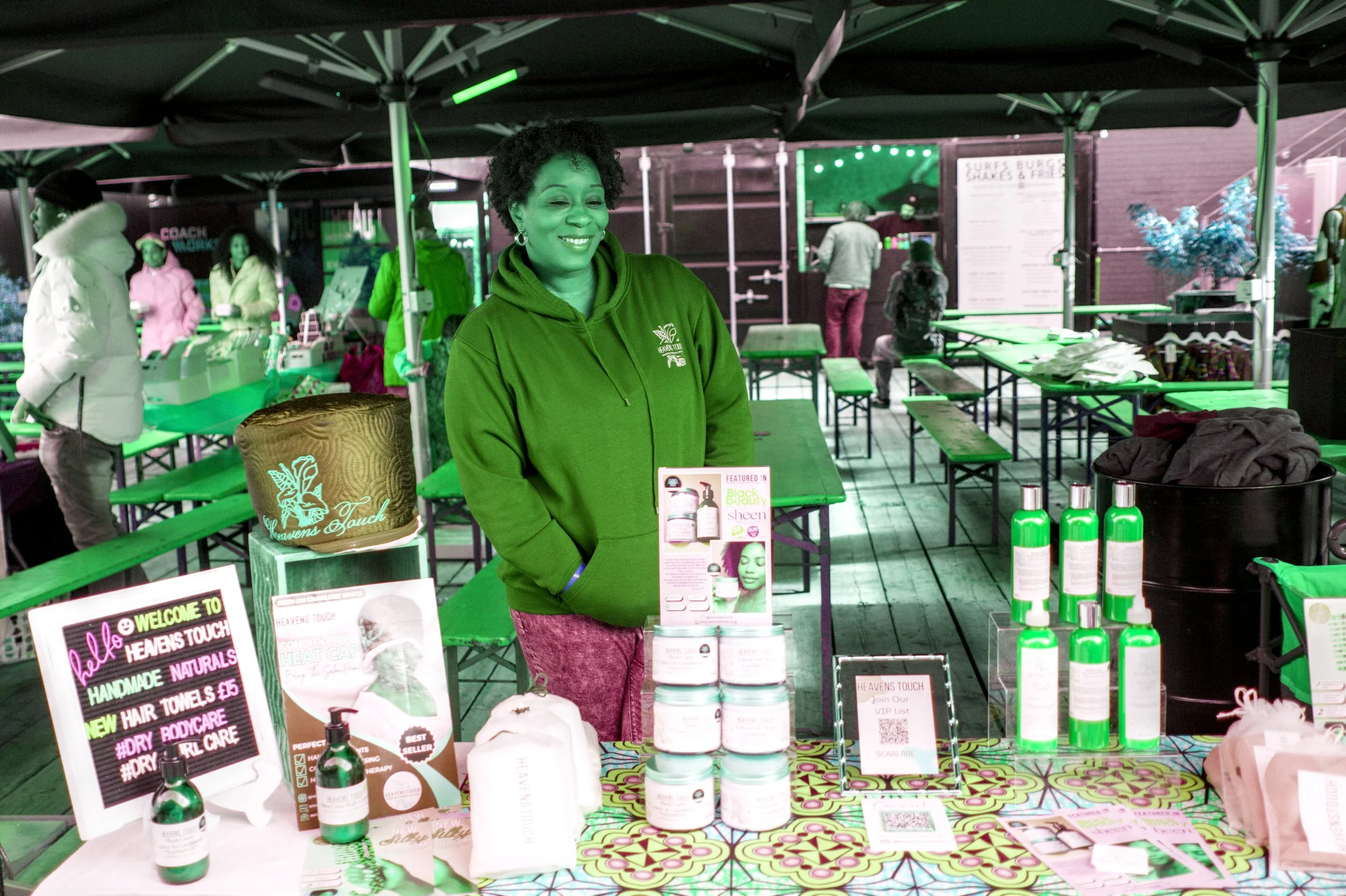 A woman wearing a maroon hoodie standing at a booth with various skincare and hair products at an outdoor market.