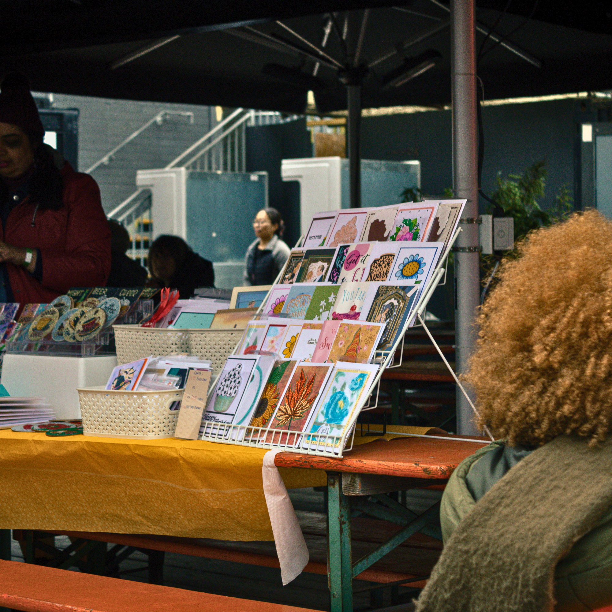 A display of greeting cards with colorful designs and messages on a metal rack at an outdoor market, with a person visible in the foreground.