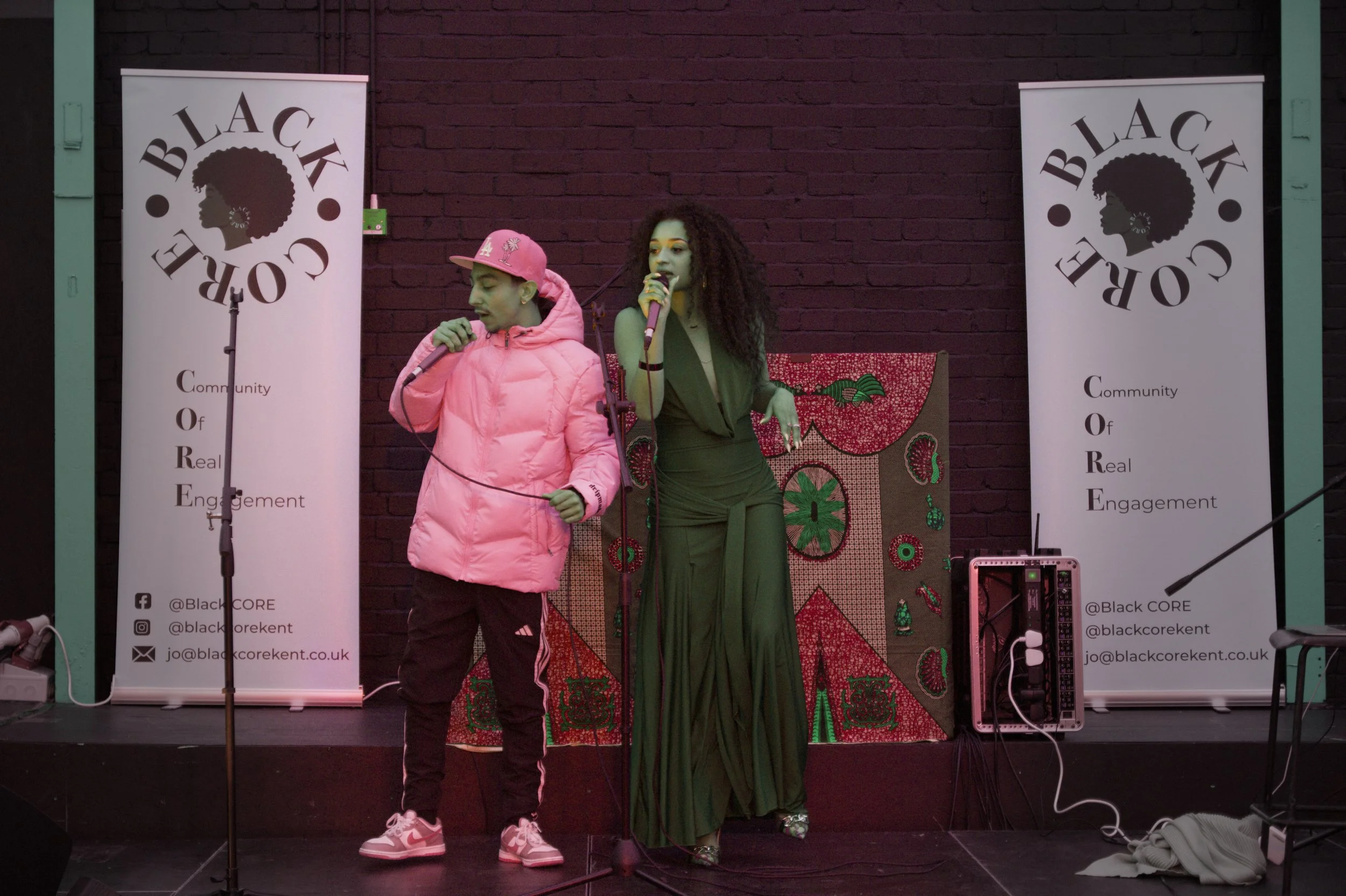 Two performers, a young man with a gray LA baseball cap and a white jacket, and a woman with long curly hair in a brown dress, are singing into microphones on a stage with a black brick background. There are two white banners with the Black CORE logo and contact information on either side of the stage.