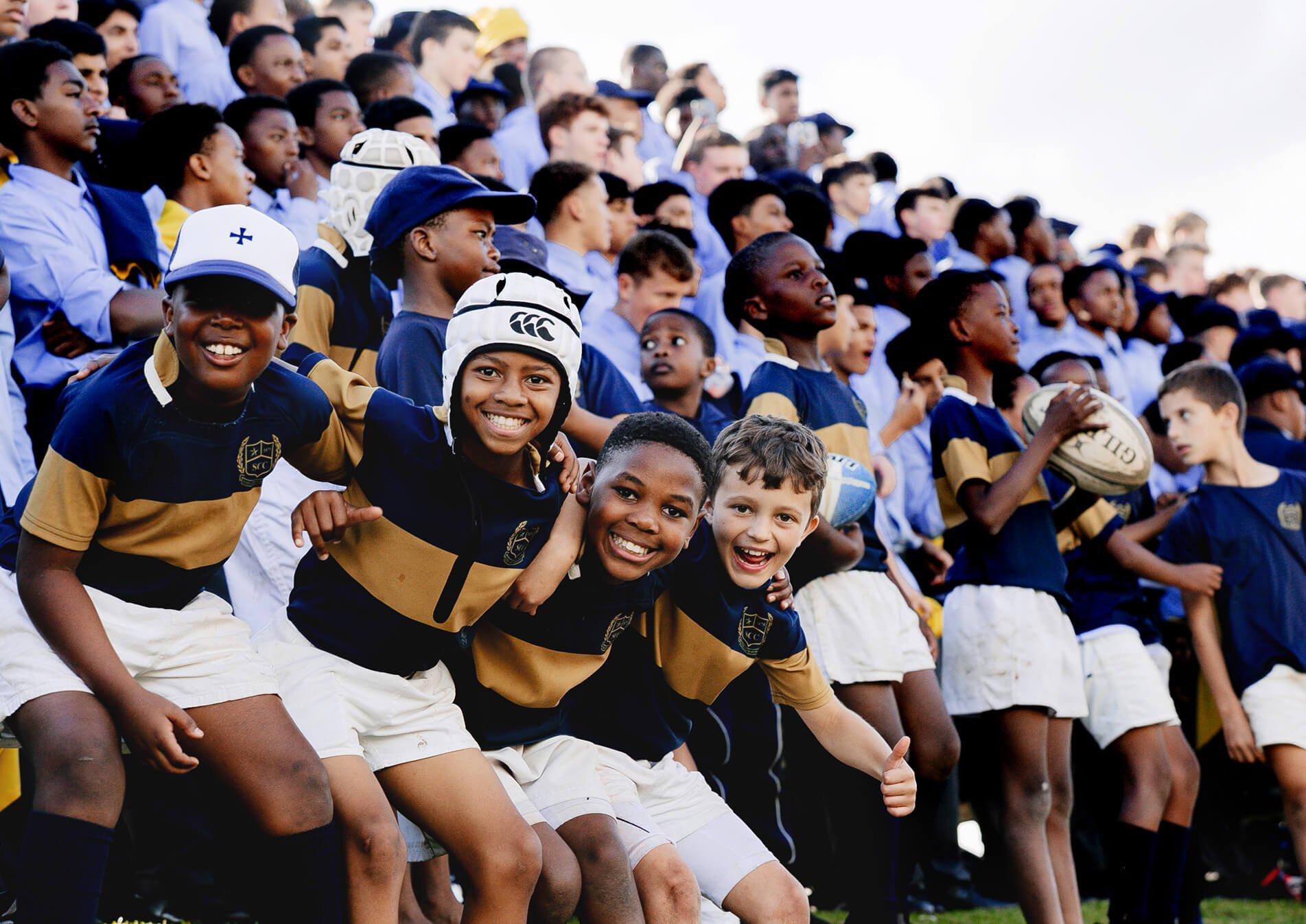 Children on bleachers at a sports event, some holding rugby balls, wearing navy and gold uniforms, smiling and cheering.