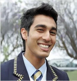 Young man in a school uniform smiling outdoors with trees in the background.