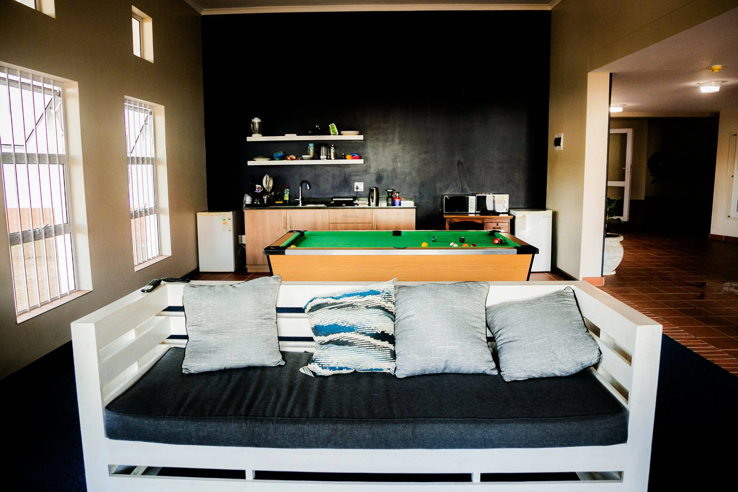 Living room with a couch, pillows, windows with bars, a black accent wall, a small kitchenette with shelves, a pool table, and some kitchen appliances.