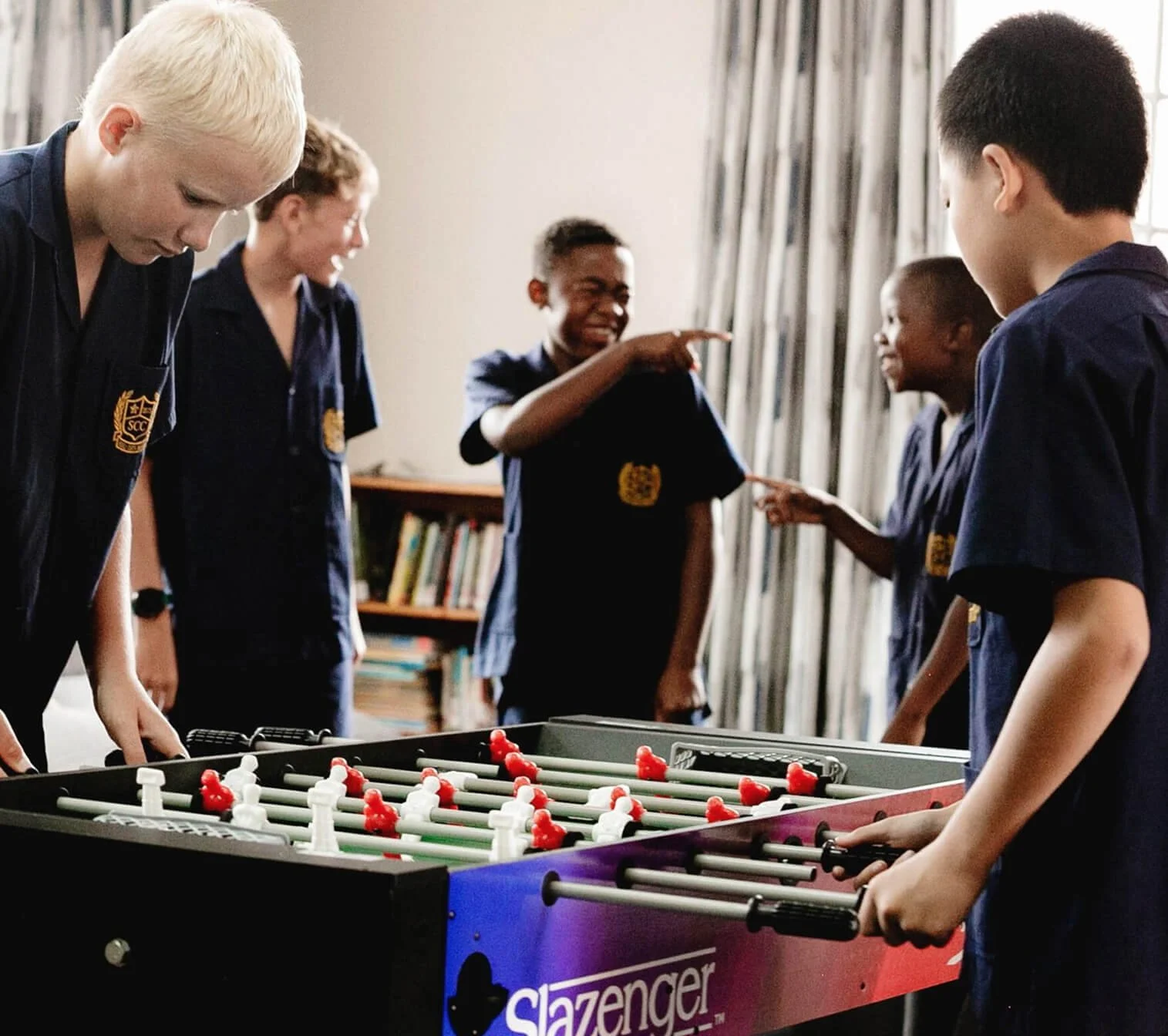 Children playing table soccer in a room with bookshelves and curtains, some smiling and engaging in conversation.