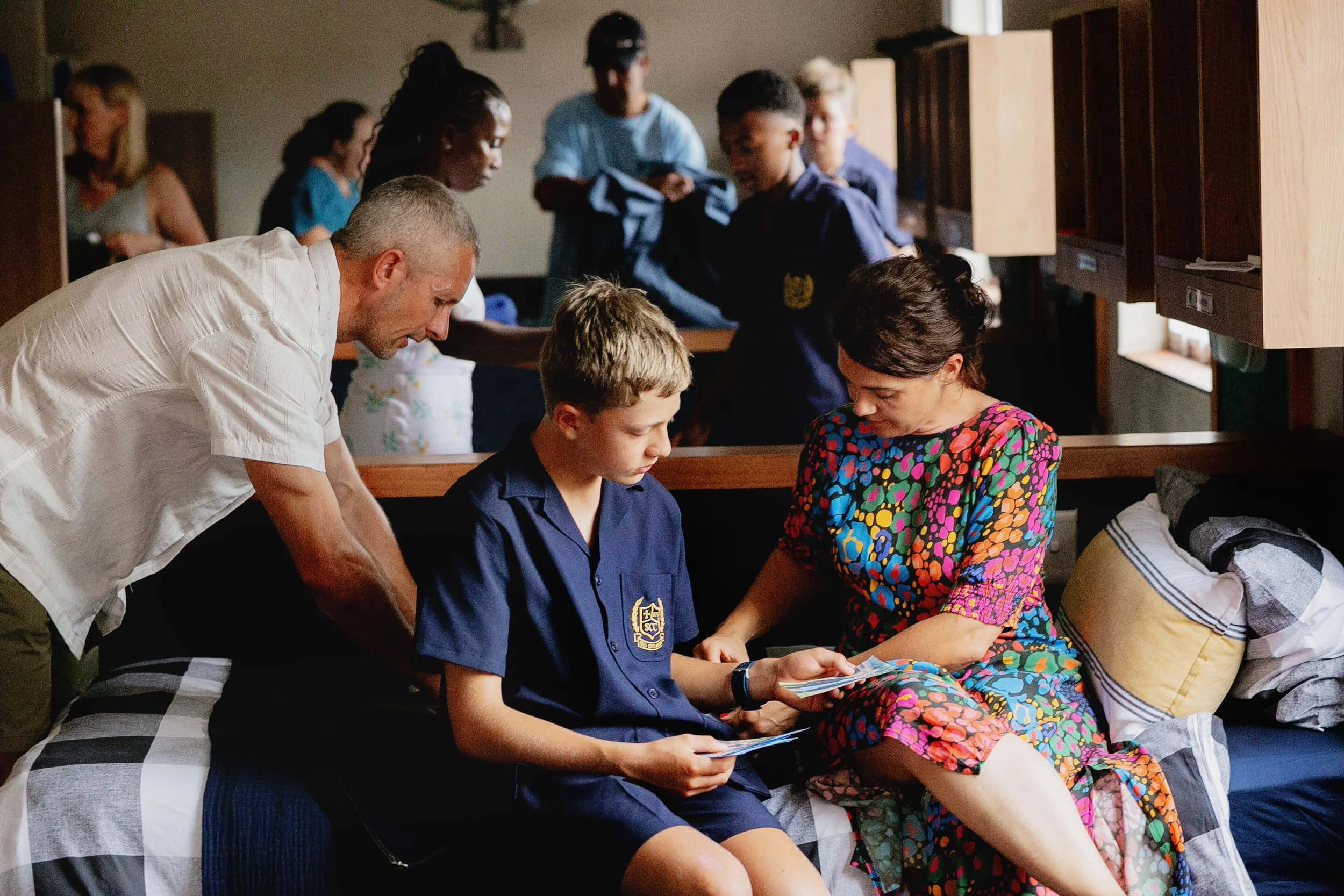 A group of people, including children and adults, are gathered in a room, sitting or standing close together, some looking at papers or a book, inside a cozy indoor setting with wooden furniture and natural light.