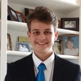 A young man in a formal suit and blue tie smiling in a room with white shelves and family photos in the background.