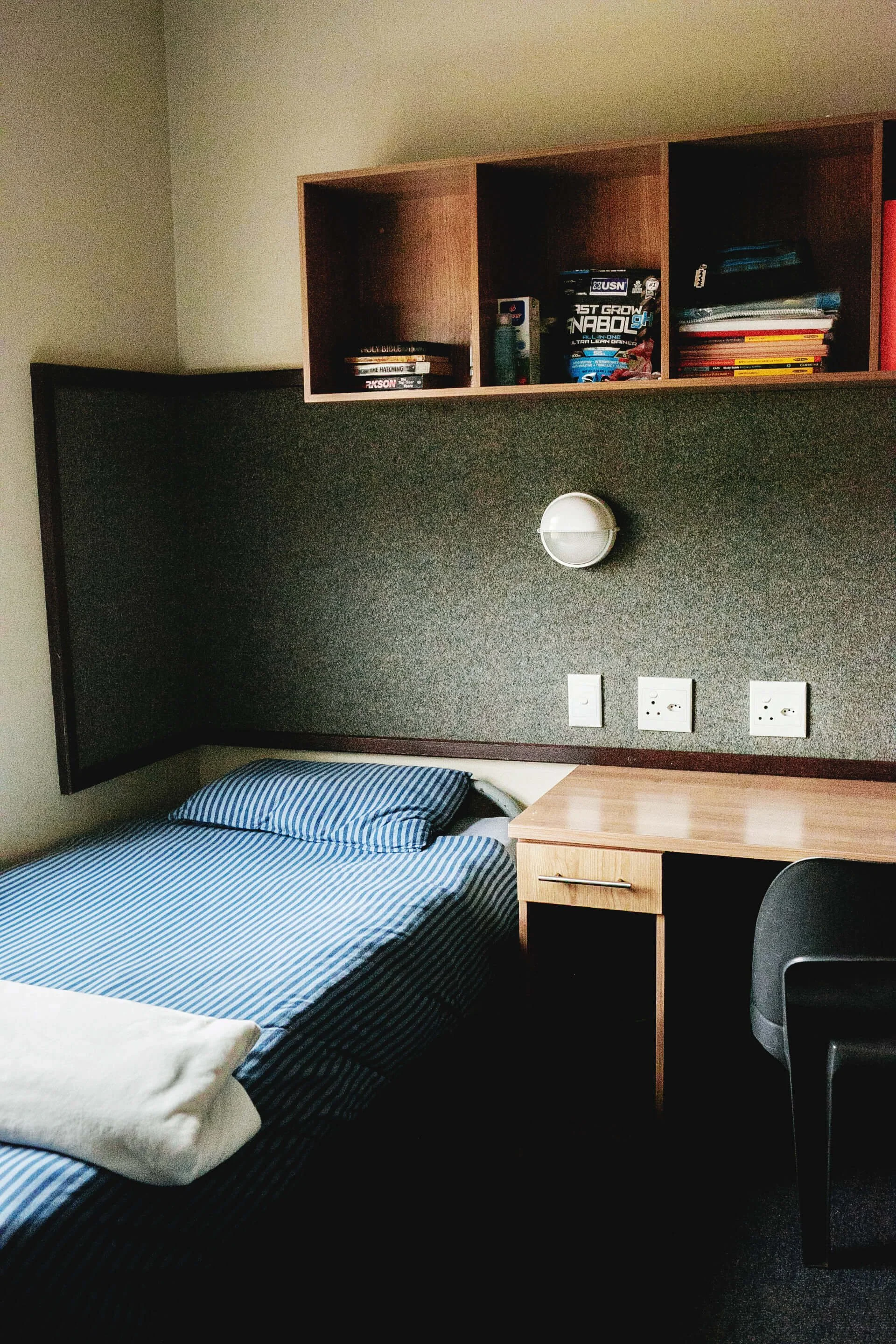 A bedroom with a bed featuring black and blue striped sheets, a pillow, and a blanket. There is a wooden nightstand with a handle next to the bed. The wall behind has a dark textured panel with three electrical outlets. Above the panel, a wall-mounted light fixture is visible. A wooden shelf with books, magazines, and a box is mounted on the wall above the panel.