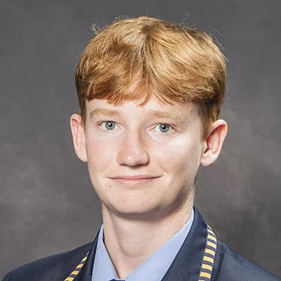 Portrait of a young man with red hair, wearing a dark blazer with a blue shirt, smiling mildly against a gray background.