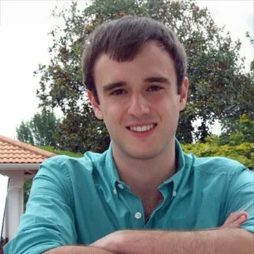 Young man with short dark hair smiling outdoors, wearing a turquoise button-up shirt, with trees and a house in the background.