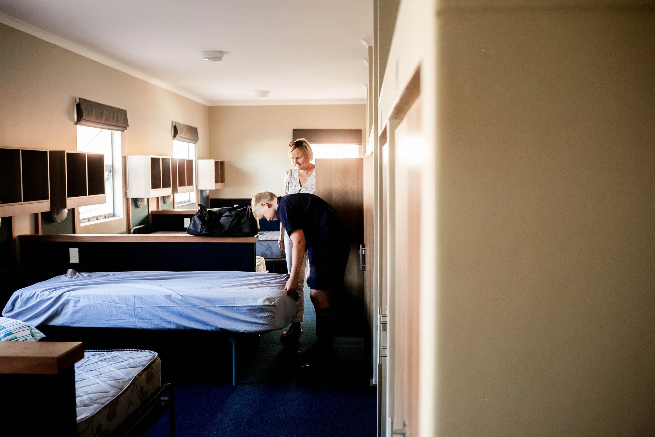 A woman and a young boy make the bed in a hotel room with beige walls, dark blue carpet, and wooden furniture.