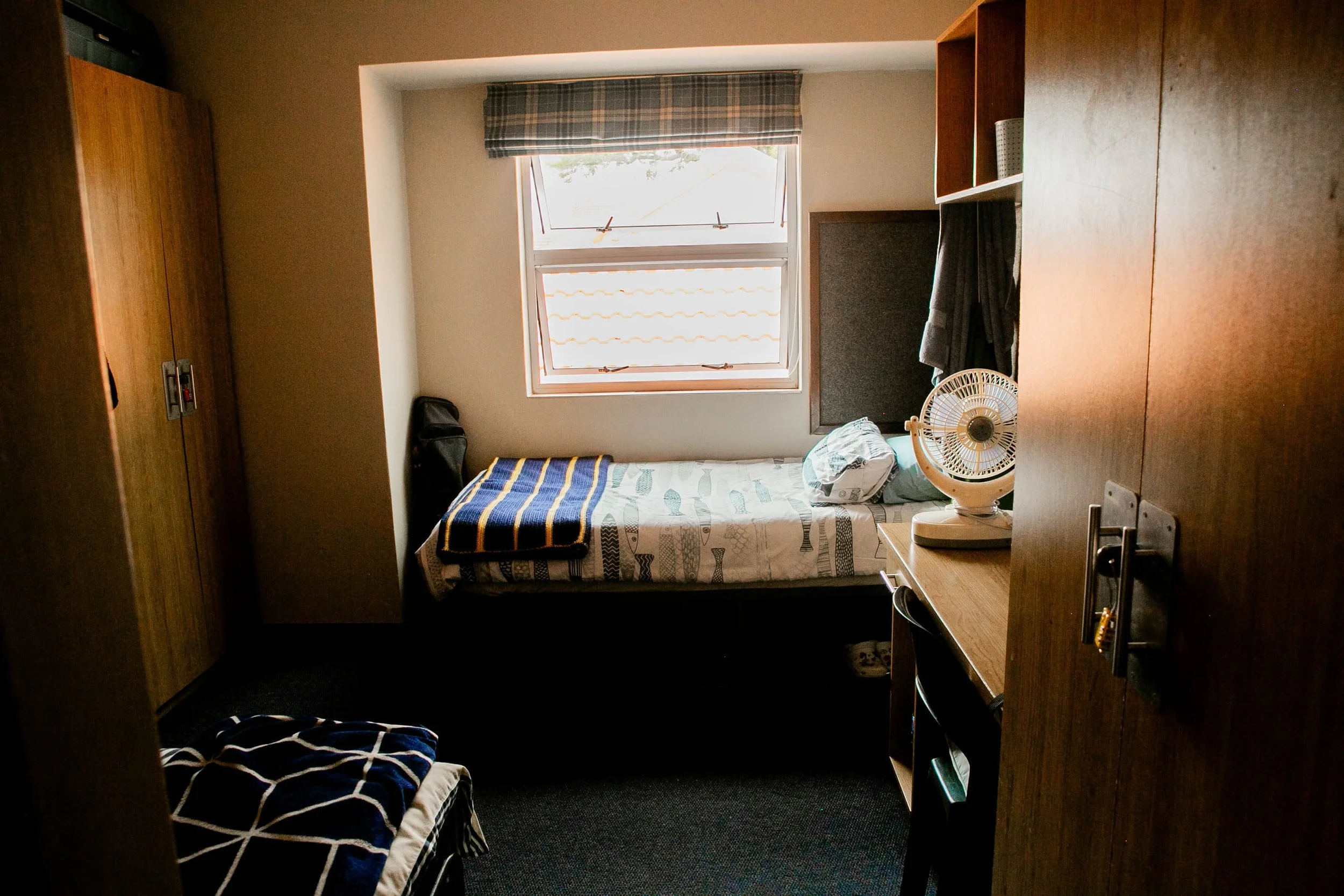 A small bedroom with a single bed in front of a window, with a checkered blanket and pillow. There is a fan on a bedside table, a wardrobe, and a wooden desk on the right side. The window has a plaid curtain.