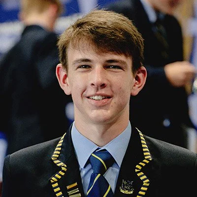 A young man in a suit and tie smiling at the camera, with a blurred background of other people in formal attire.
