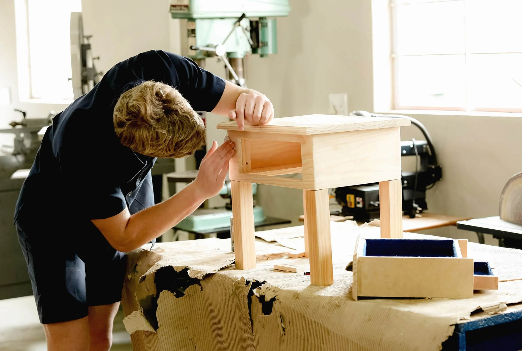 A person is sanding wood furniture in a workshop, using a block sander with a wooden table in the background.