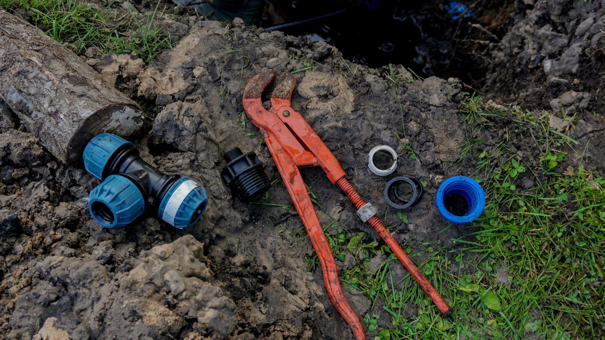 Tools and pipe fittings on the ground near a trench for plumbing work, including a red pipe wrench, several blue and black pipe connectors, and a section of pipe.