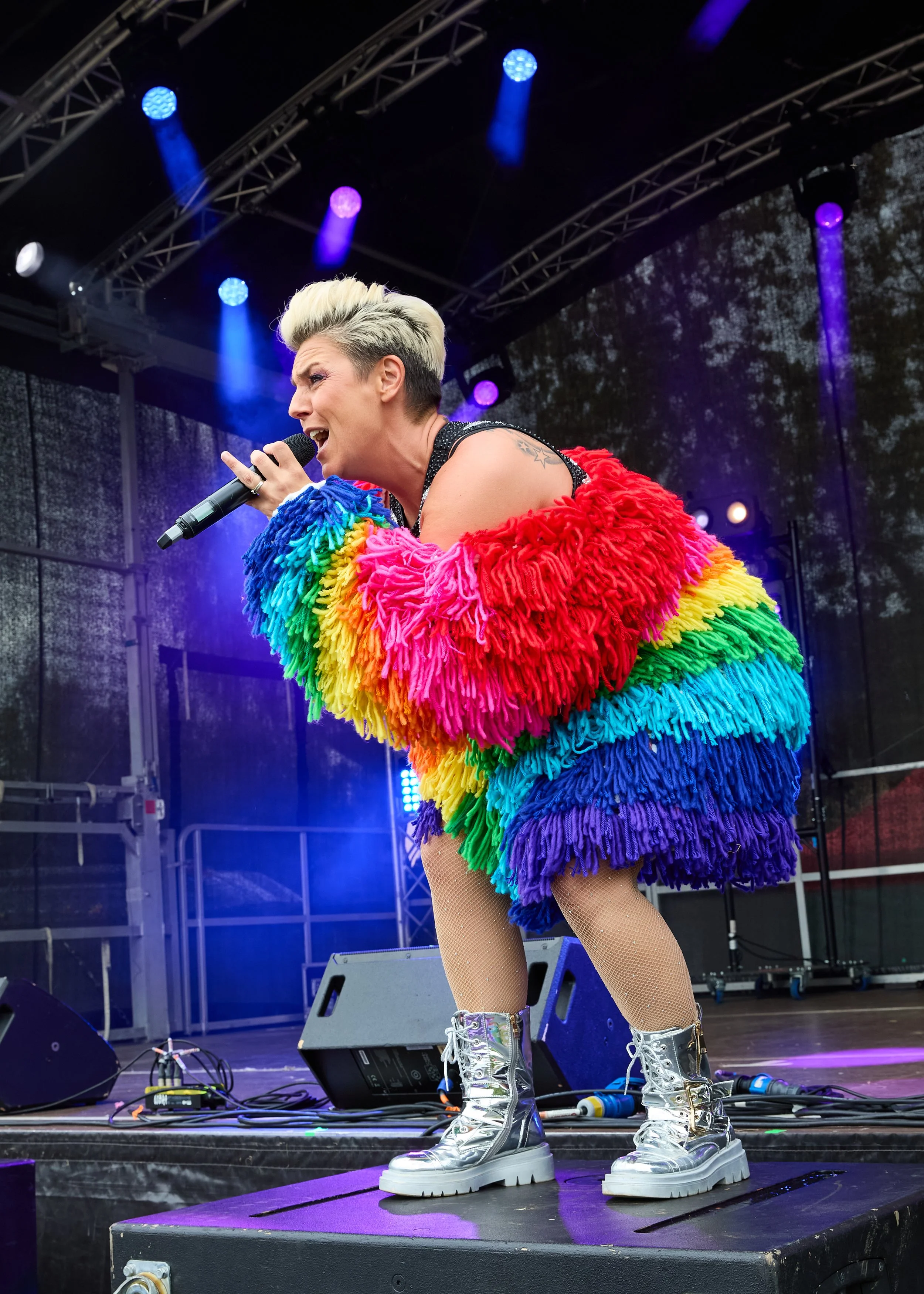 Performer singing on stage wearing a rainbow-colored fluffy jacket, silver boots, and fishnet stockings, with stage lights overhead.