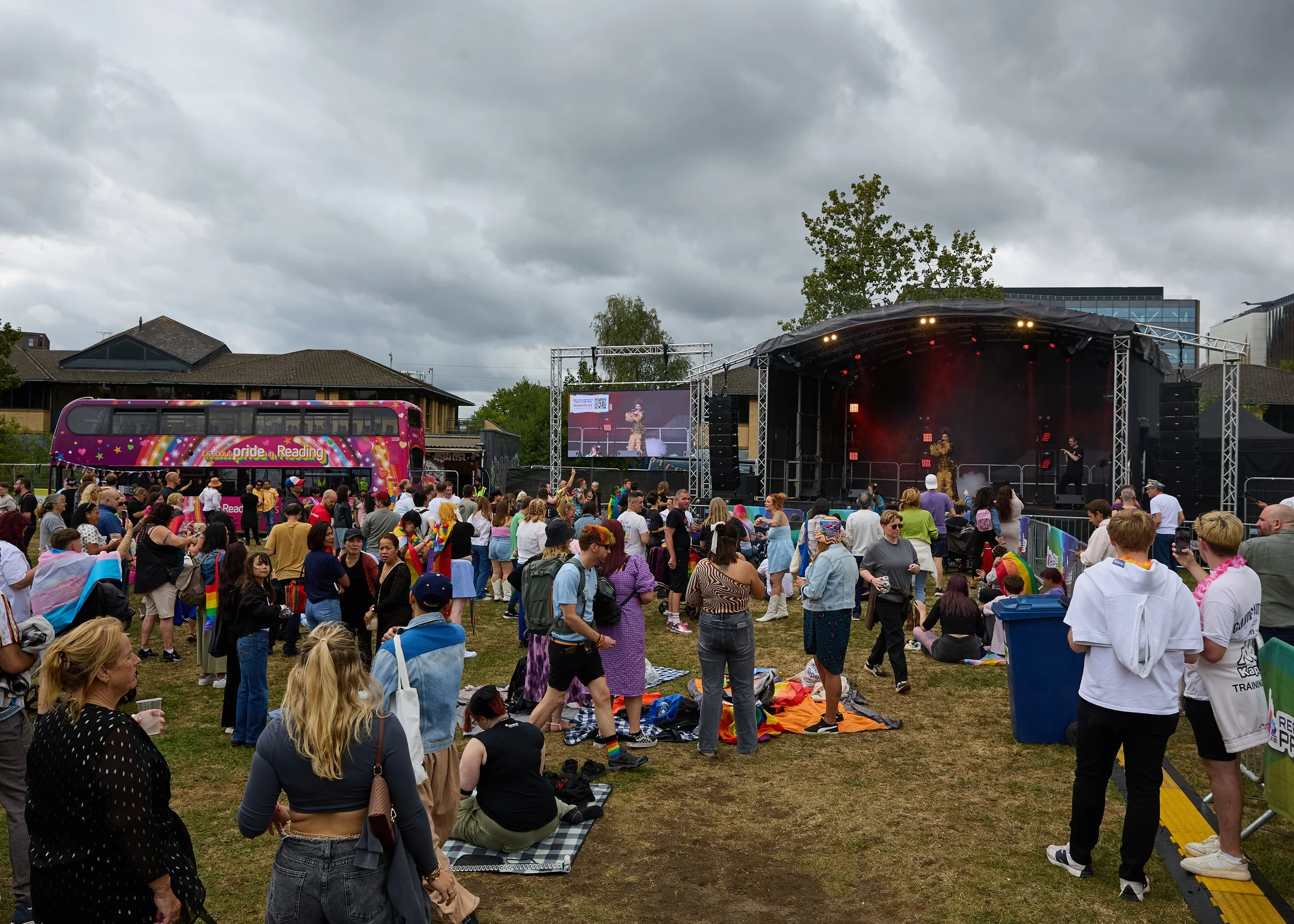 Crowd of people gathered outdoors for a pride event with a stage and large screen, overcast sky, rainbow flags, and a pink double-decker bus decorated with pride colors and the text 'It's about pride in Reading'.