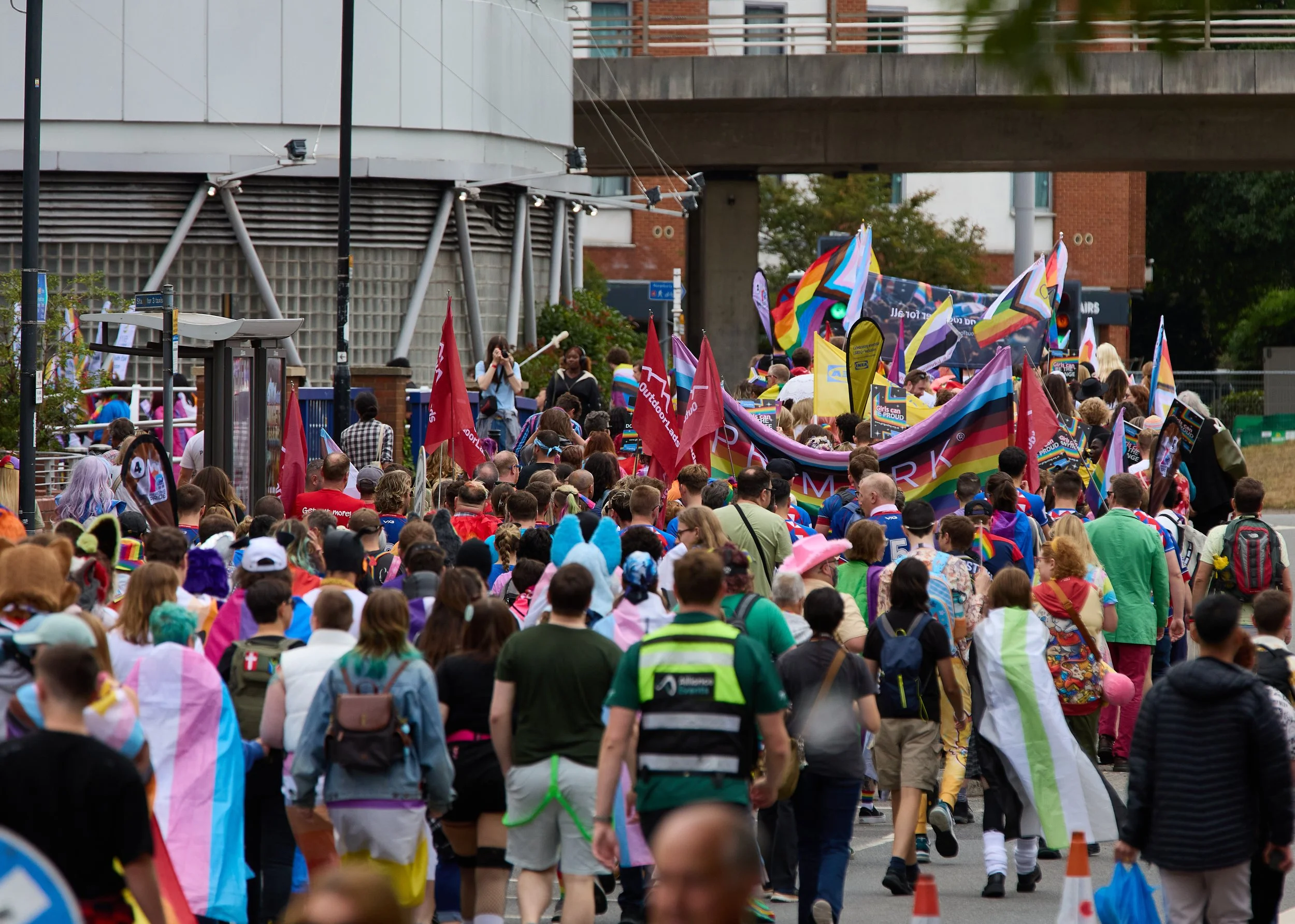 Crowd of people participating in a pride parade with rainbow flags, banners, and colorful attire in an urban outdoor setting.