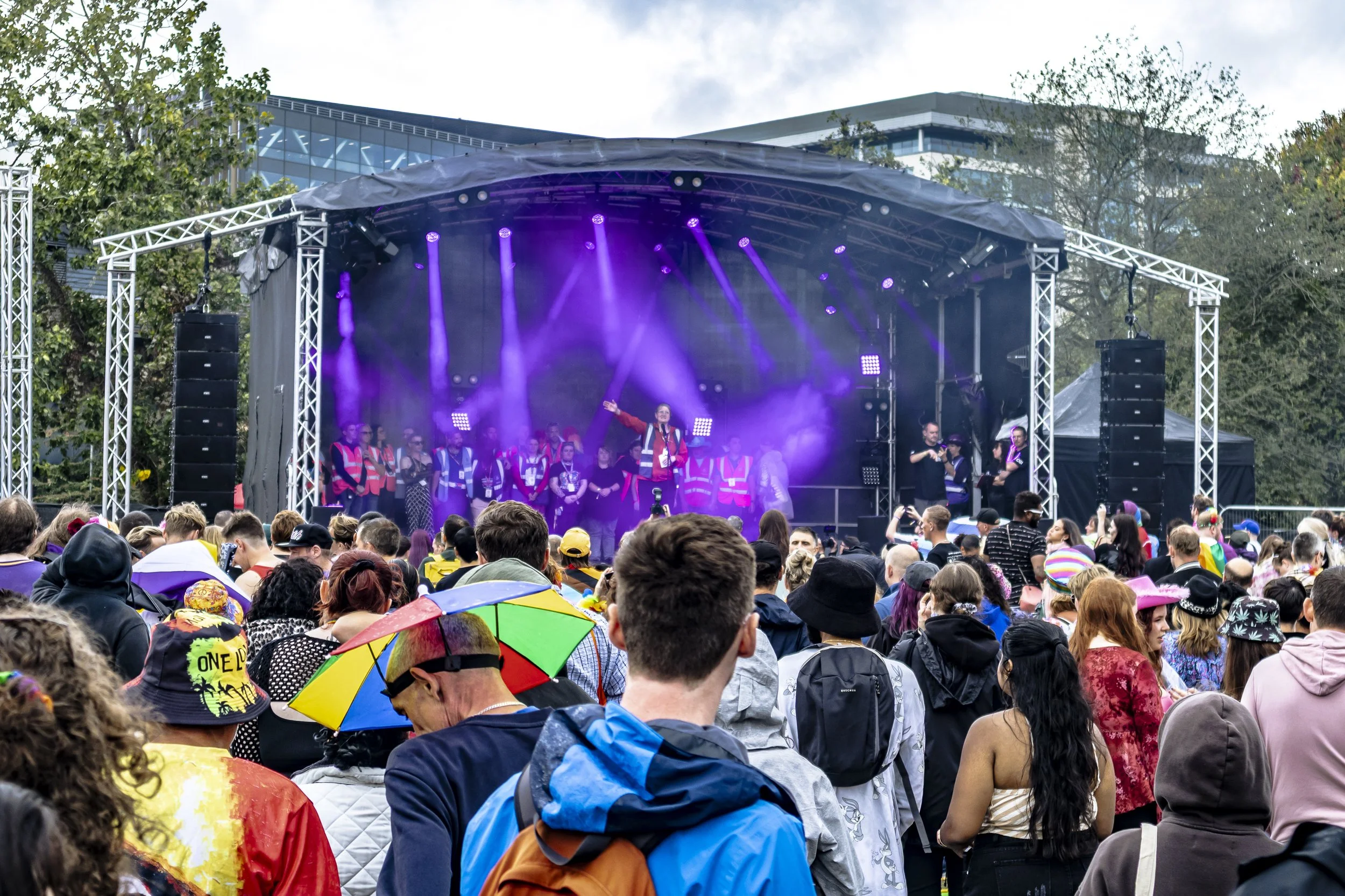 Crowd of people attending an outdoor event or concert with a stage, purple lighting, and a performer speaking into a microphone. Some attendees are holding colorful umbrellas.