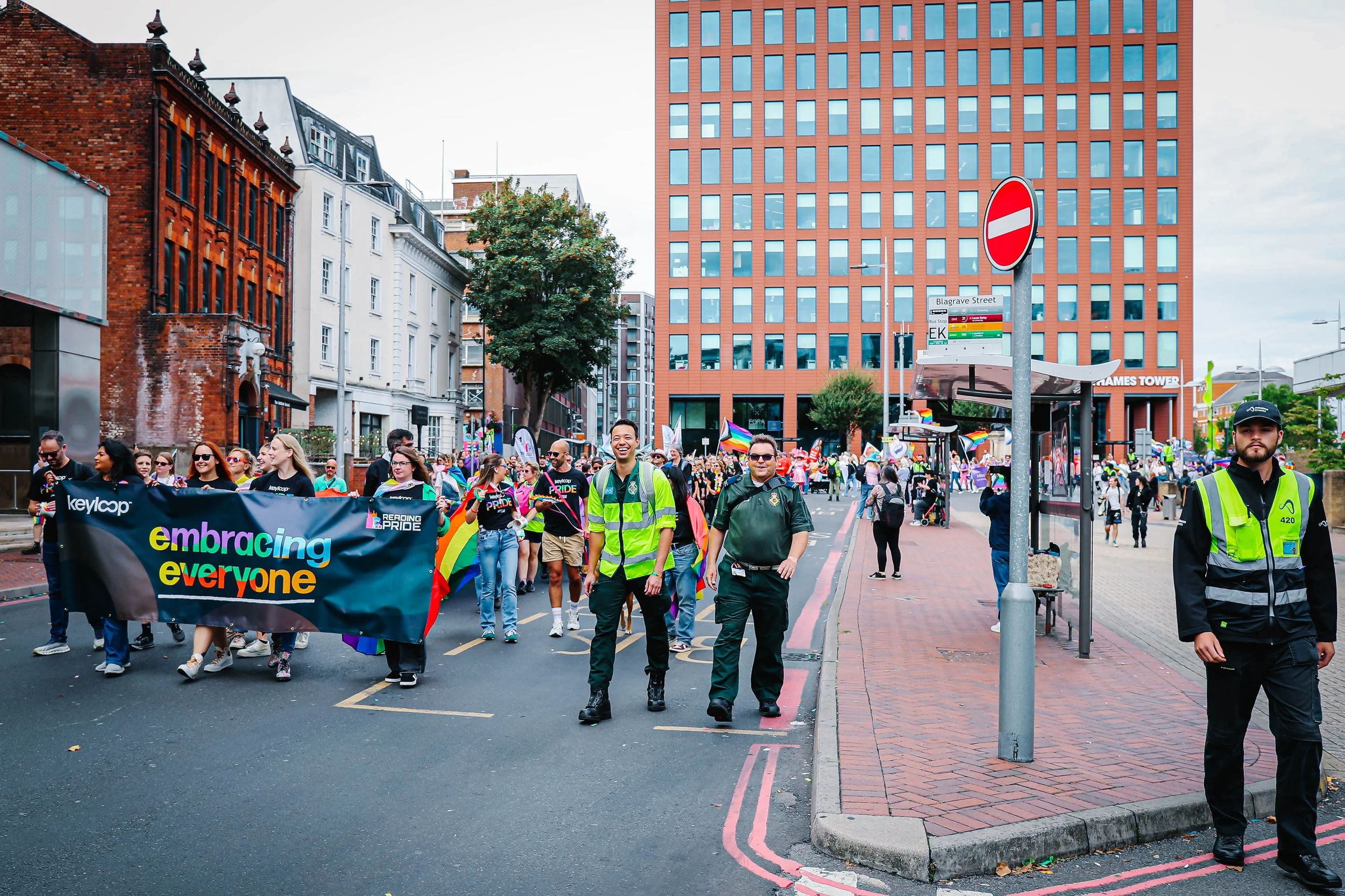 Peaceful city street during Pride parade with diverse crowd holding a banner that reads 'embracing everyone', surrounded by modern and historic buildings, including a tall red-brick office tower and older brick structures, with a large group of people marching, some waving rainbow flags, under cloudy skies.