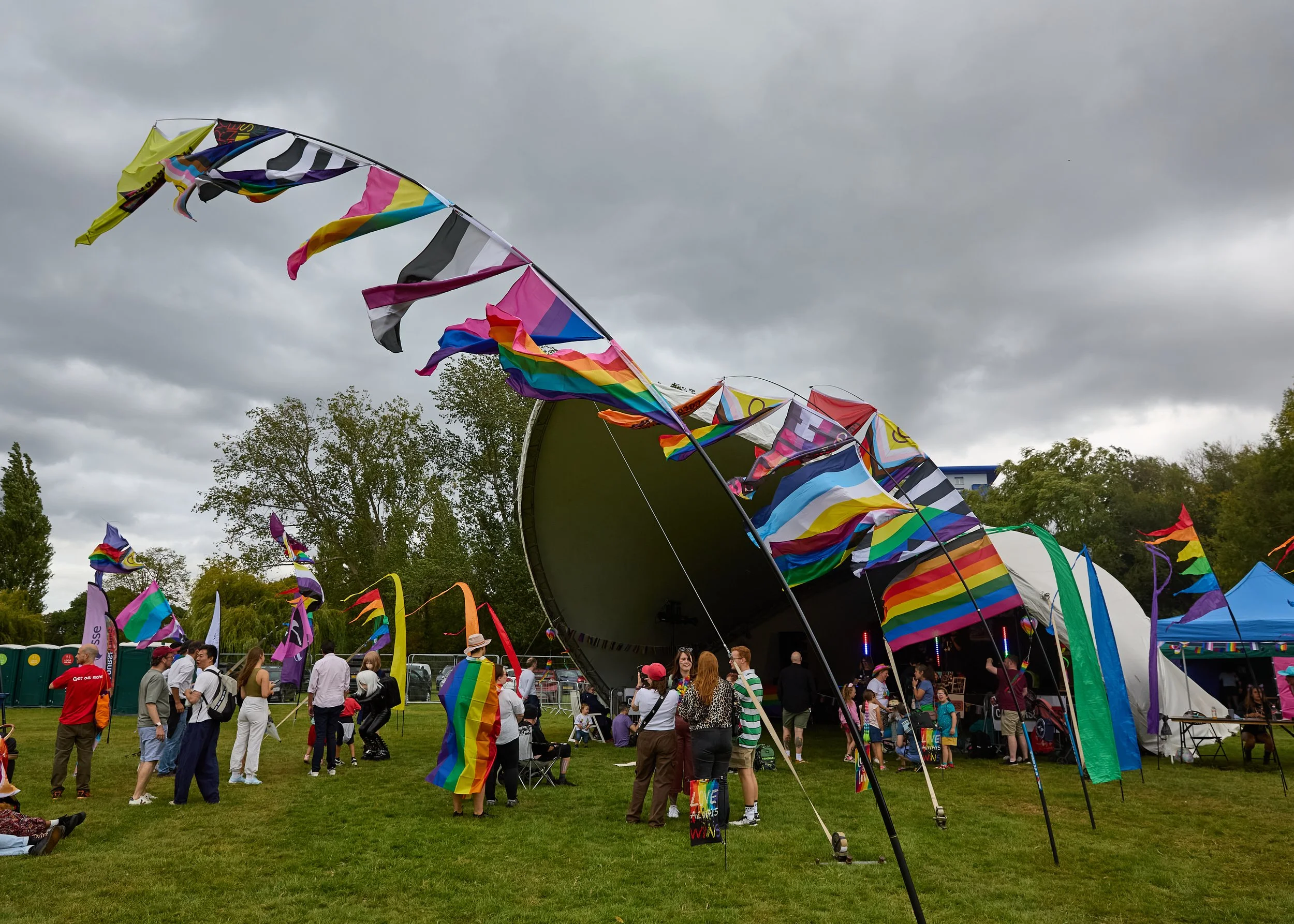 Crowd of people at an outdoor pride event with rainbow flags, tents, and banners under a cloudy sky.