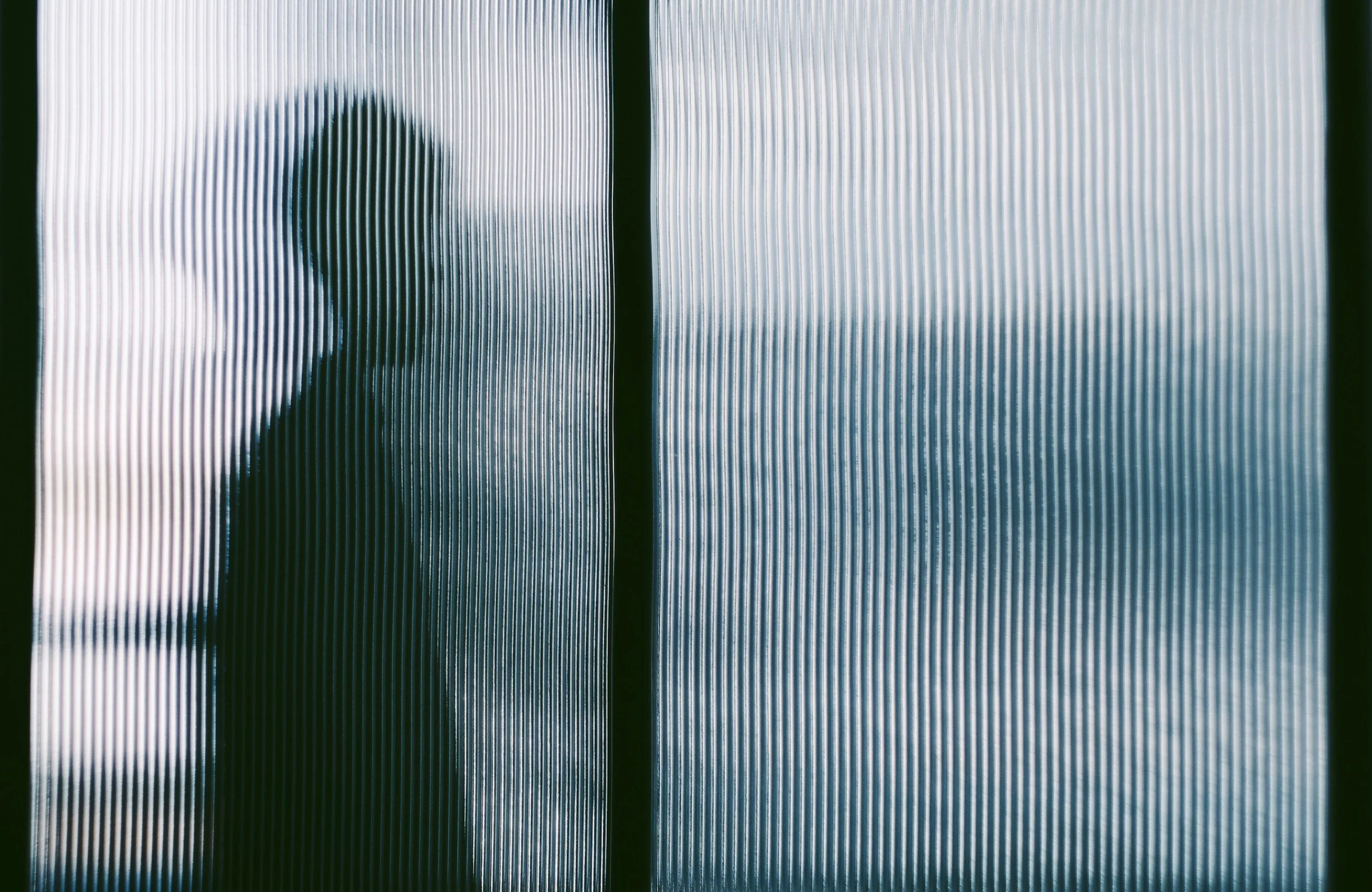 A silhouette of a person with long hair seen through textured glass with vertical ridges.
